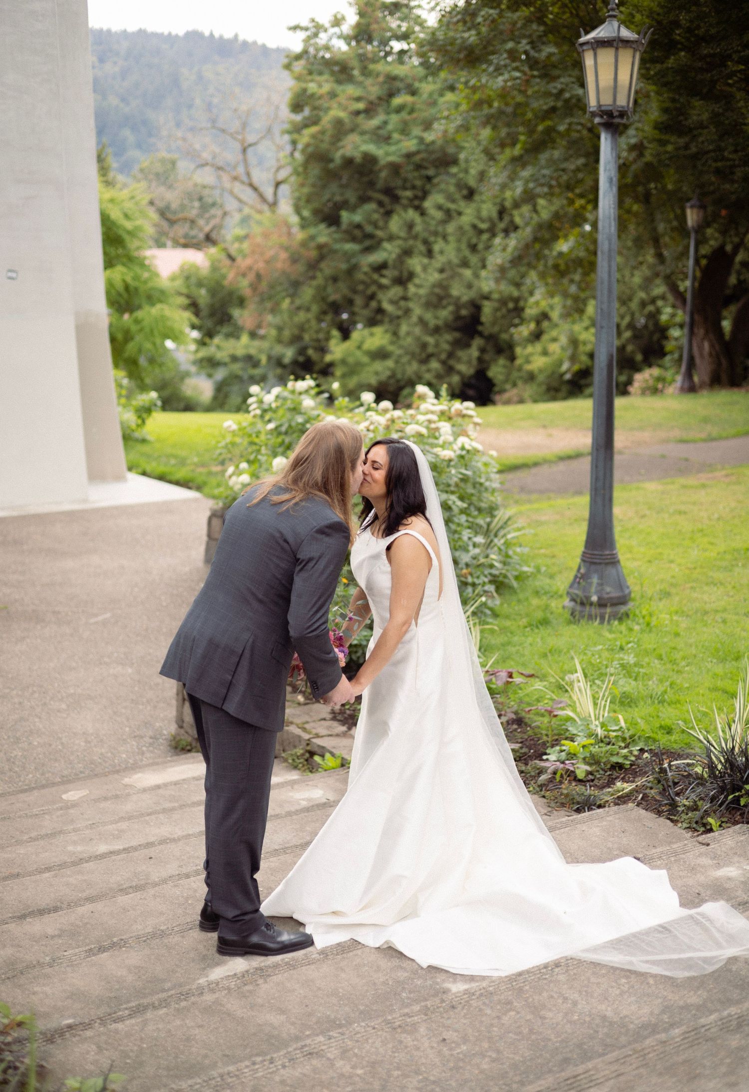 A couple shares a romantic moment on steps, bride in white gown with veil, groom in gray suit, lush greenery backdrop.