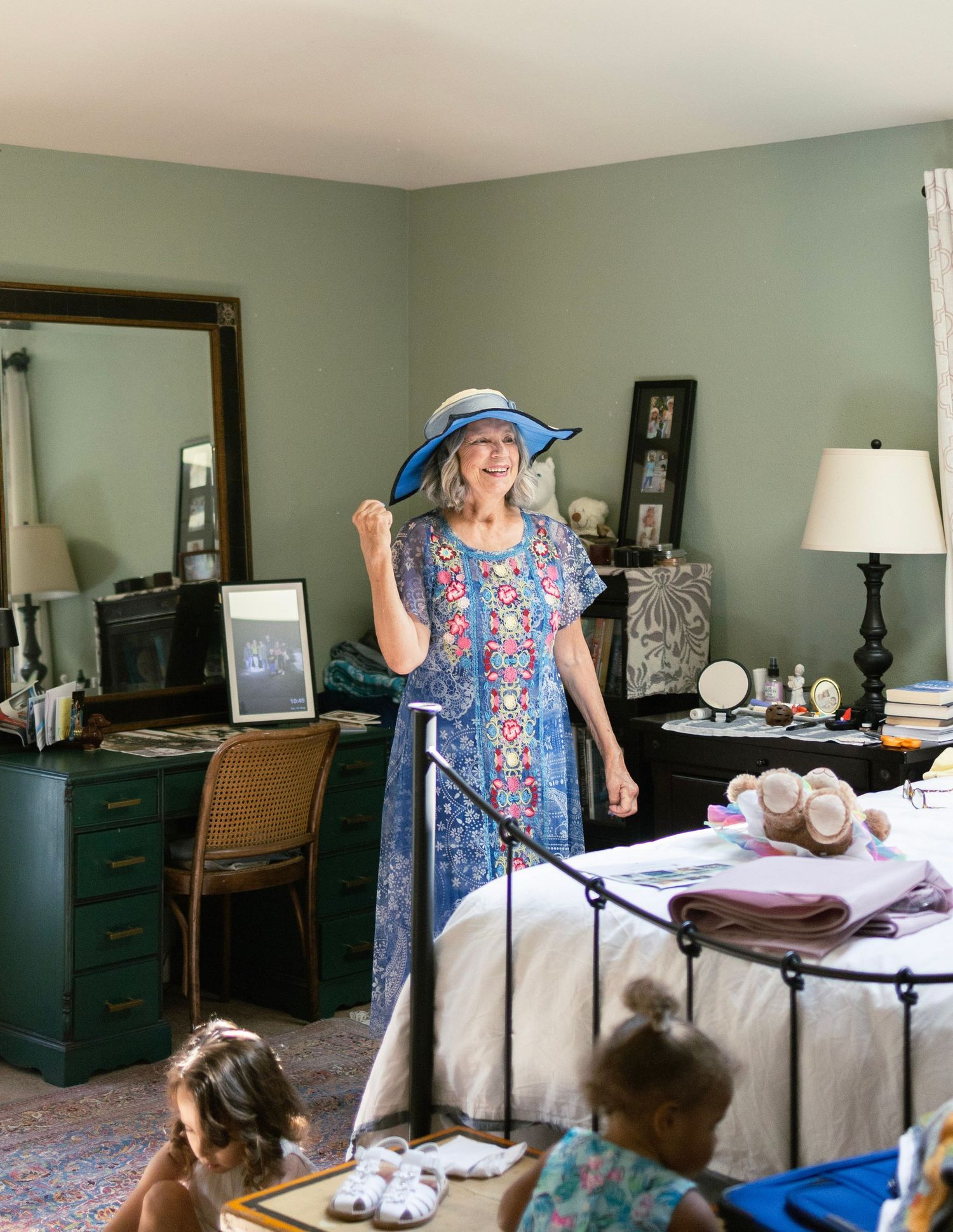 Elderly woman in blue floral dress and wide-brim hat poses cheerfully in vintage bedroom.