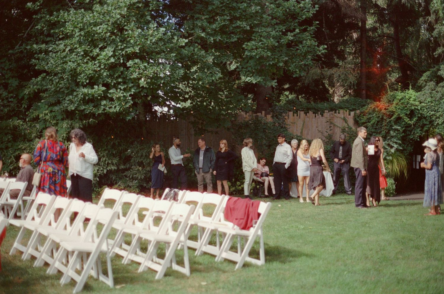 Outdoor wedding ceremony setup with white folding chairs on green lawn, guests mingling near lush trees.