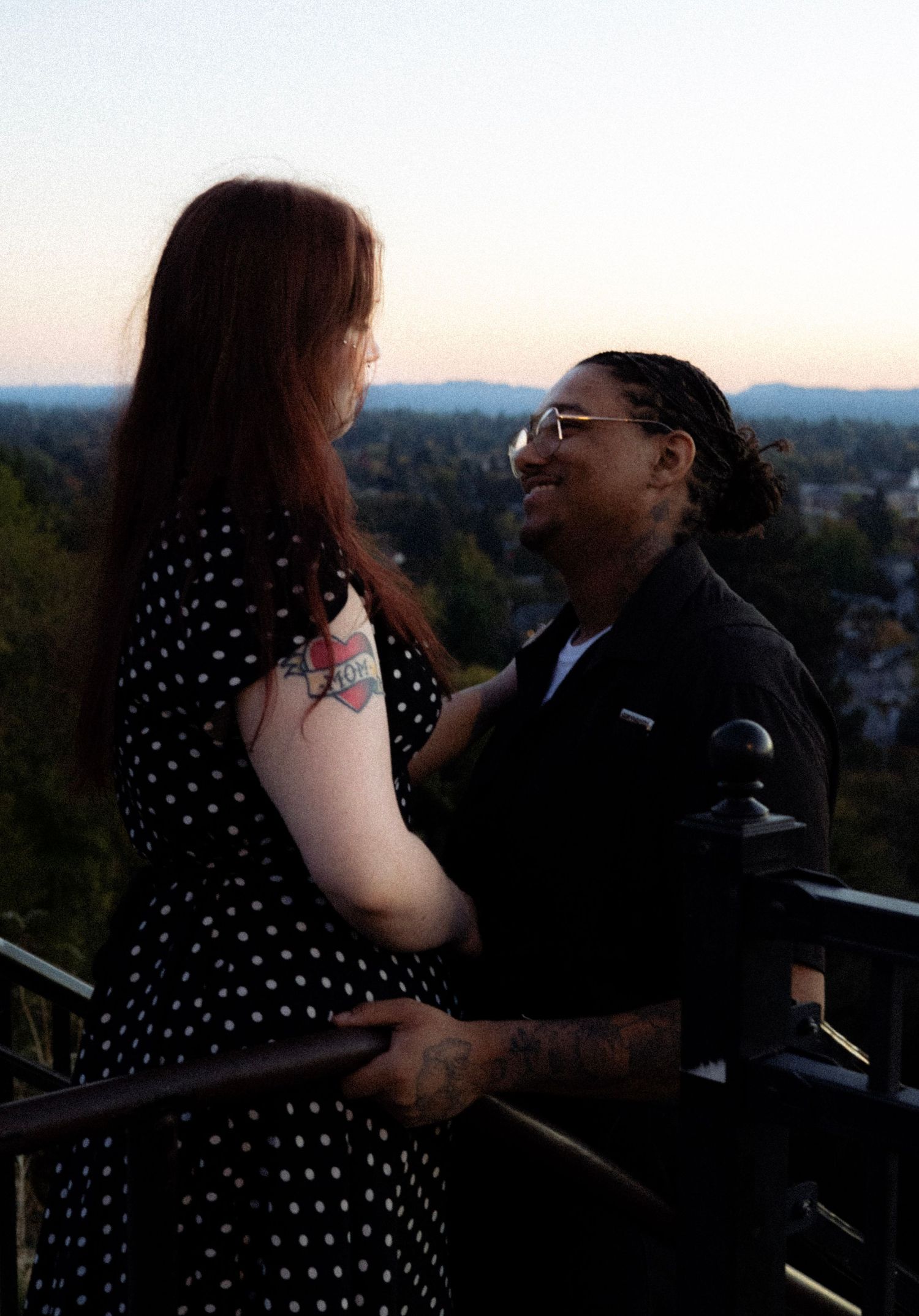 Couple sharing a romantic moment on a balcony at dusk, woman in polka dot dress with tattoo, city view behind them.