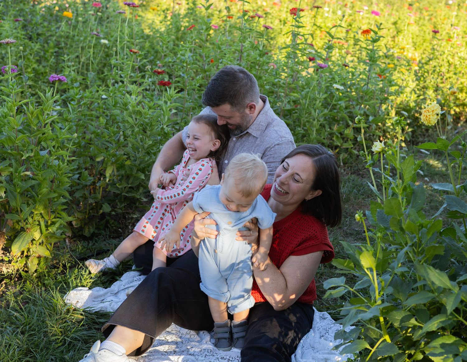 Family of four sitting in a colorful wildflower meadow, parents cuddling two young children on a white blanket.