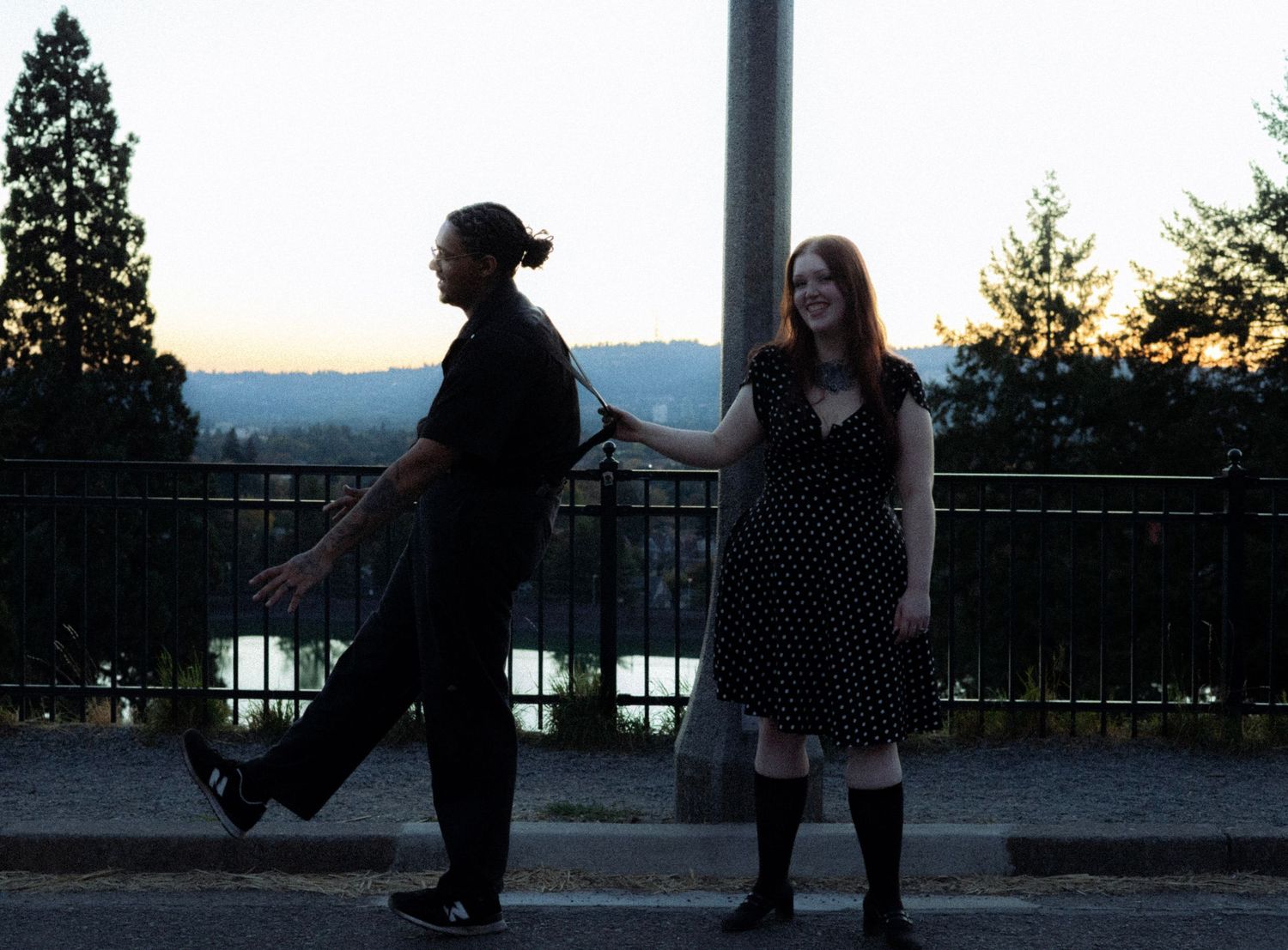A couple playfully posing at dusk near a railing with a scenic river view in the background.
