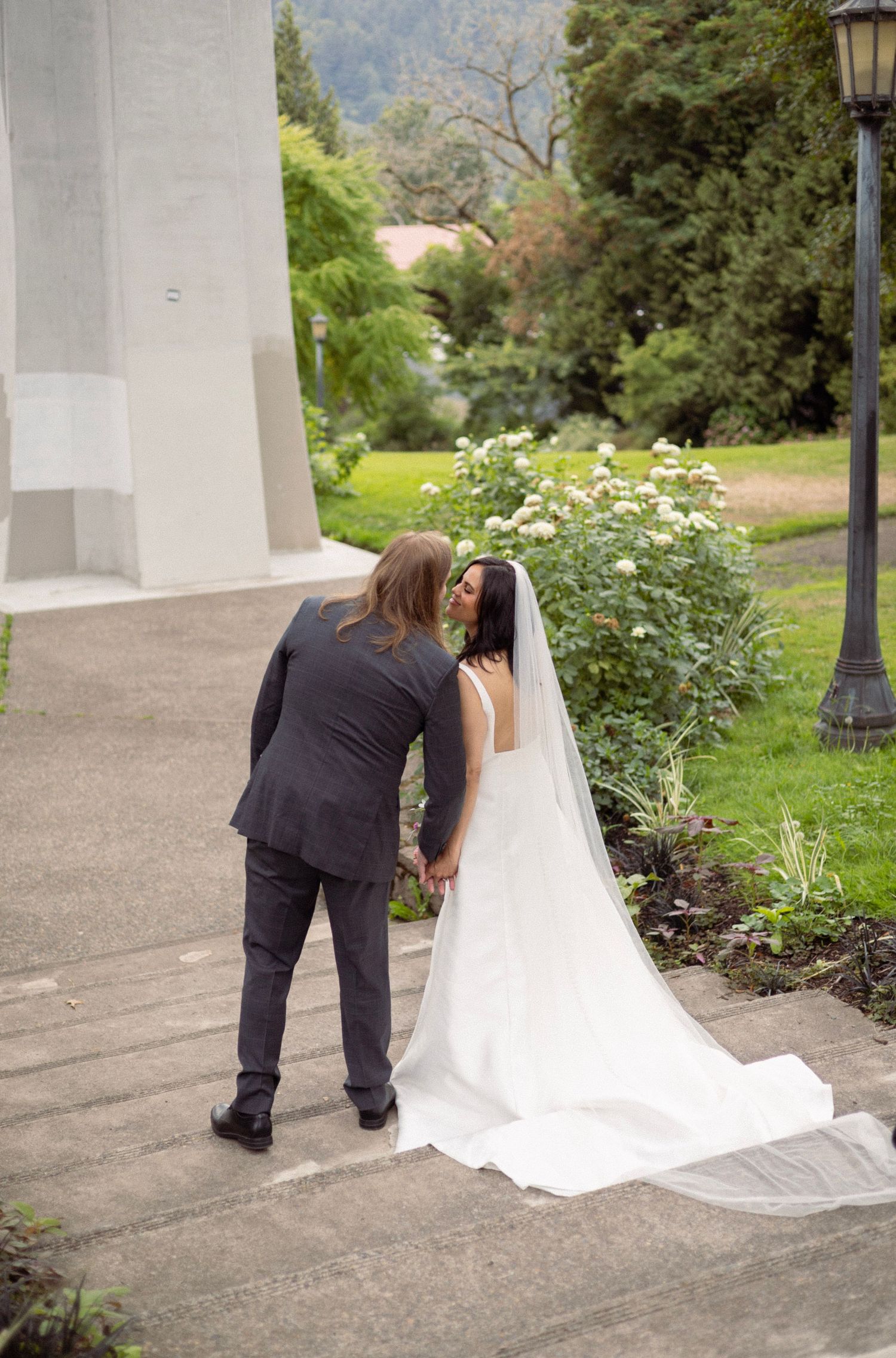 A groom in a gray suit kisses his bride in a white gown and veil in a lush garden setting.