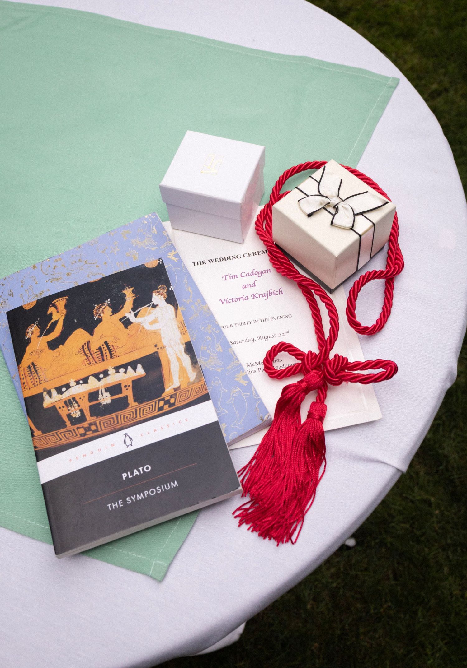 Wedding ceremony items including Platos Symposium book, ring box, red tassel cord, and ceremony program on a table.