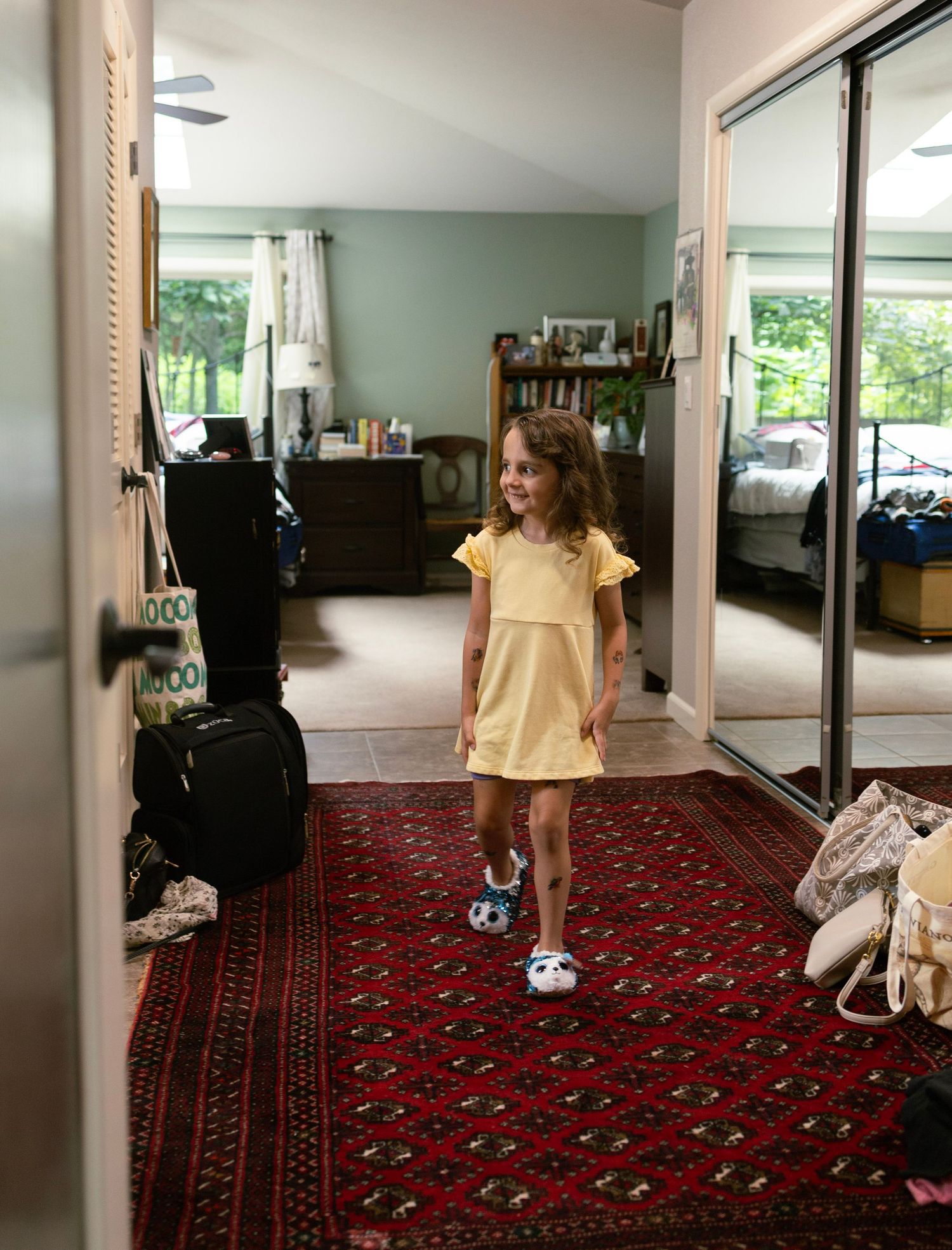 Young girl in yellow dress smiling on red rug in bedroom hallway with mirrored closet doors.