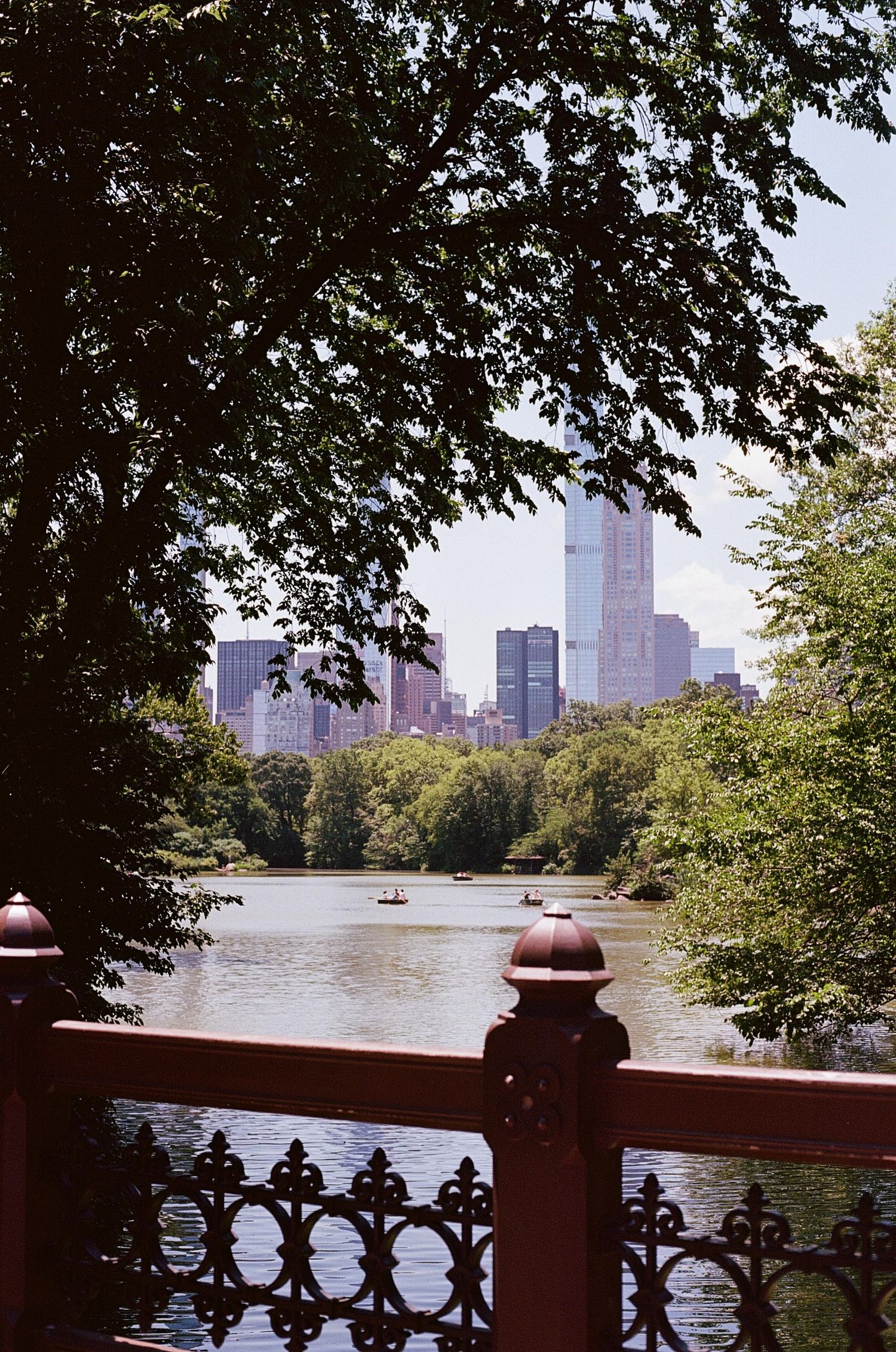 View of Central Park Lake with rowboats and Manhattan skyline visible through lush green trees from Bow Bridge.