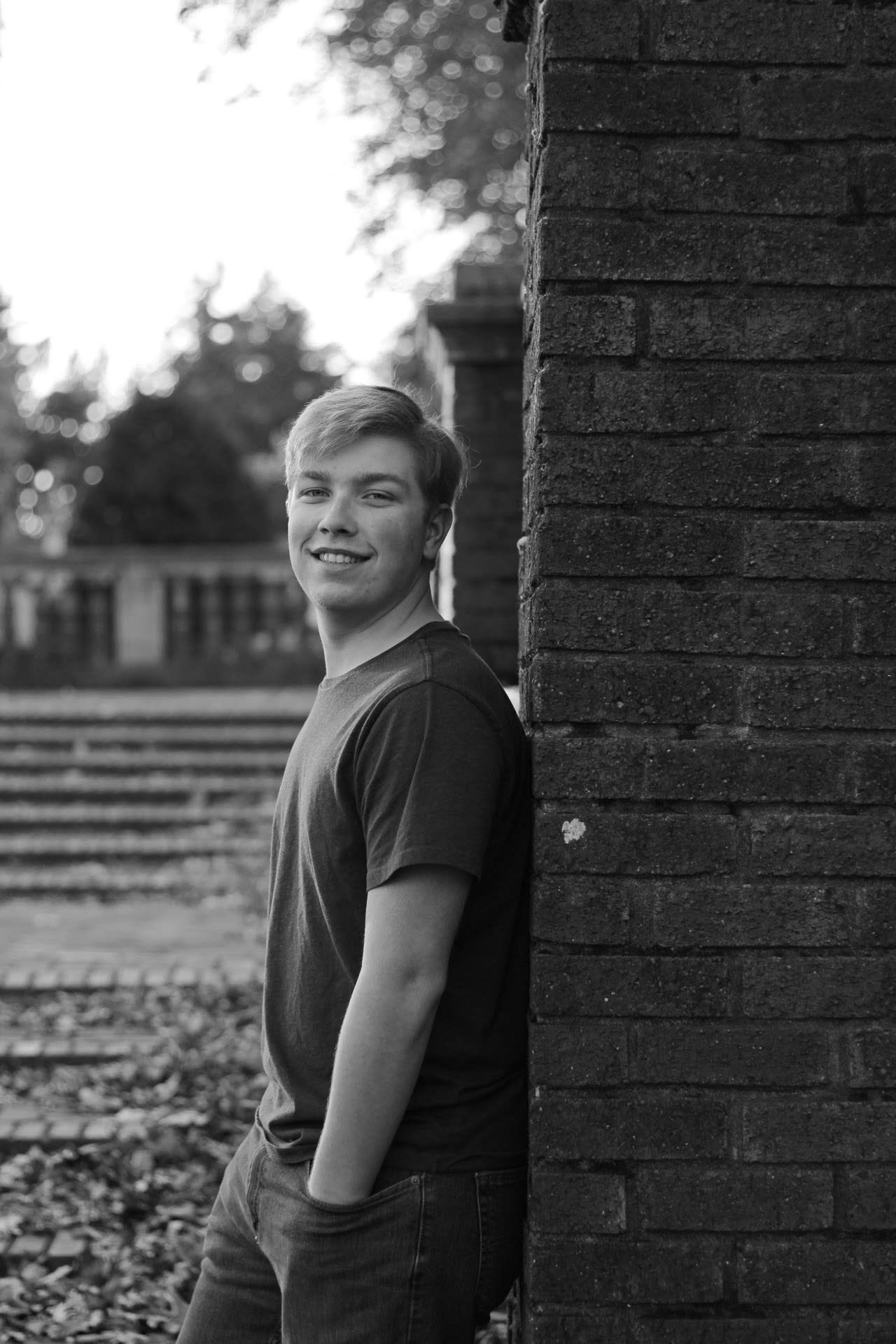 Smiling young man leaning against a brick wall outdoors in a black and white portrait photo.