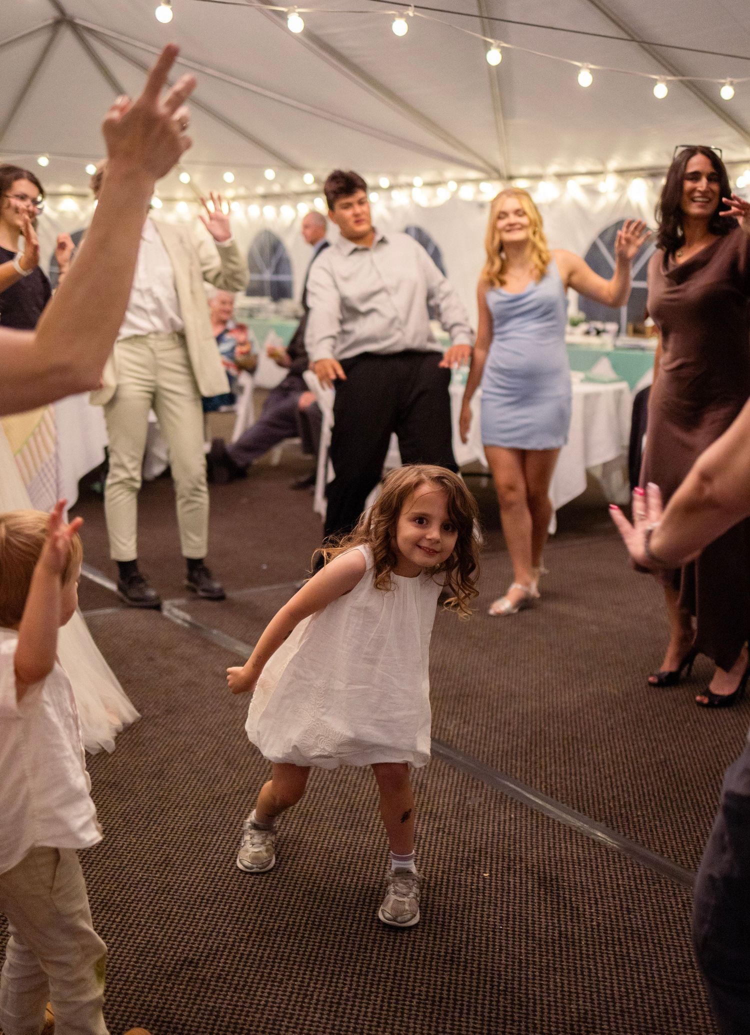 A joyful little girl in a white dress dances at a wedding reception under a tent with string lights.