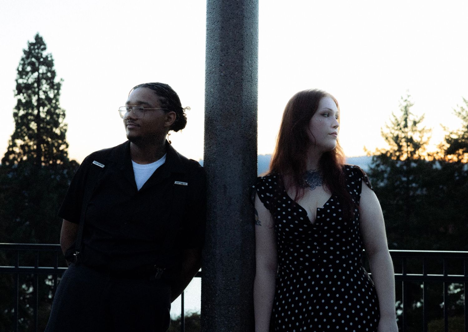 Two people standing by a pole outdoors at dusk, one in a black shirt and one in a polka dot dress.