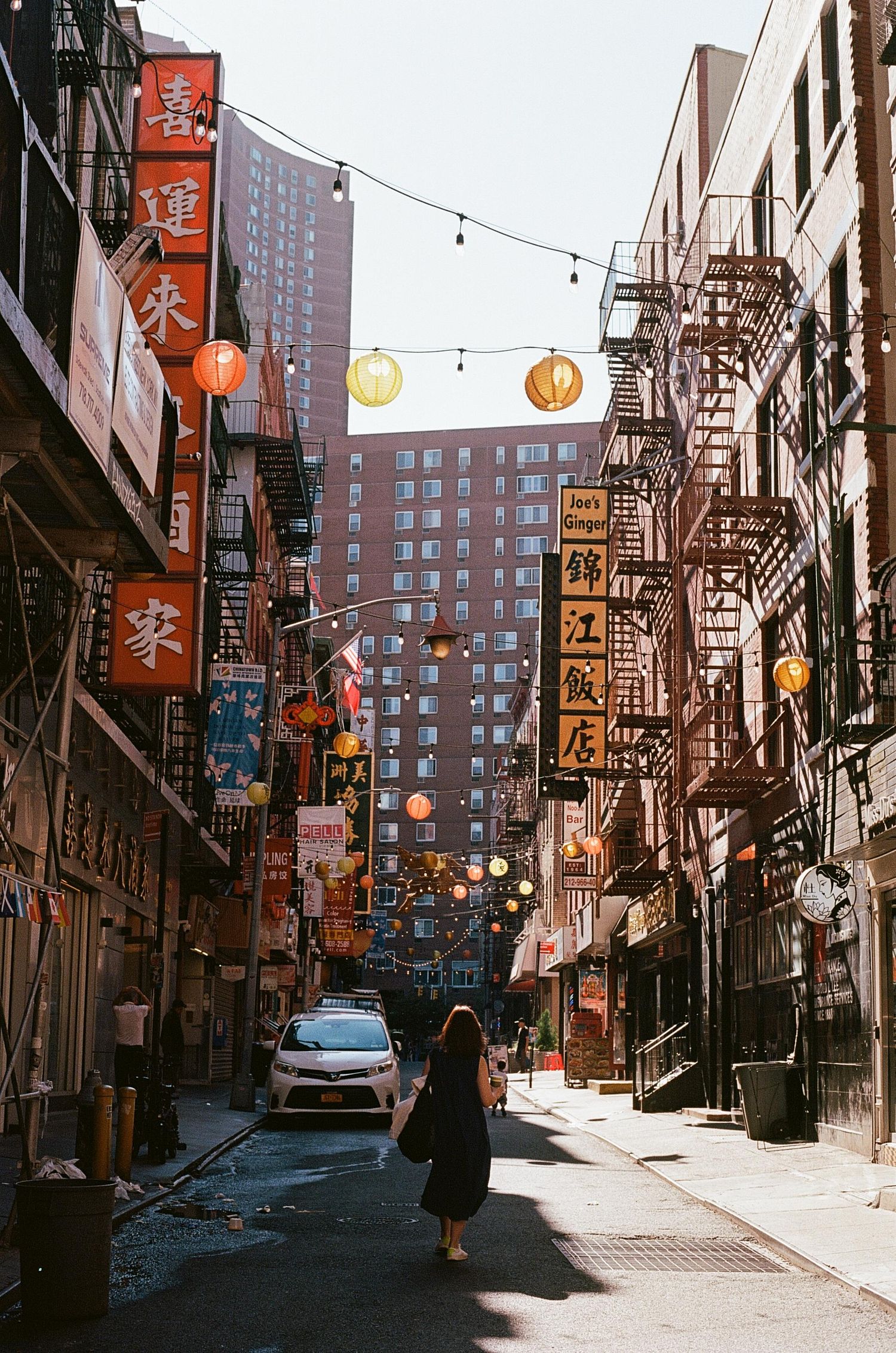 A woman walks through a narrow Chinatown alley lined with Chinese signs, lanterns, and fire escapes in NYC.