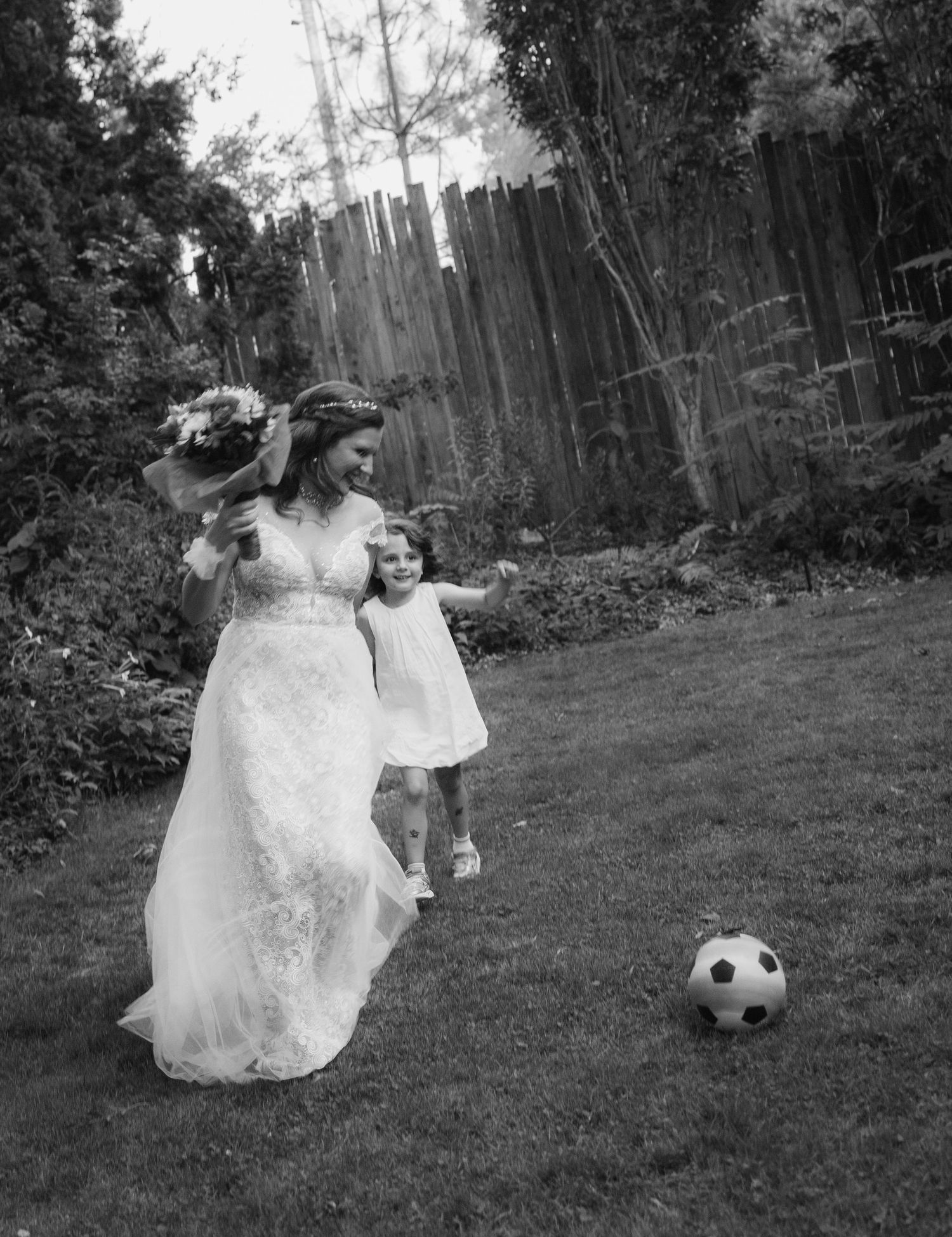 Bride in lace wedding dress plays soccer with young girl in backyard garden setting.