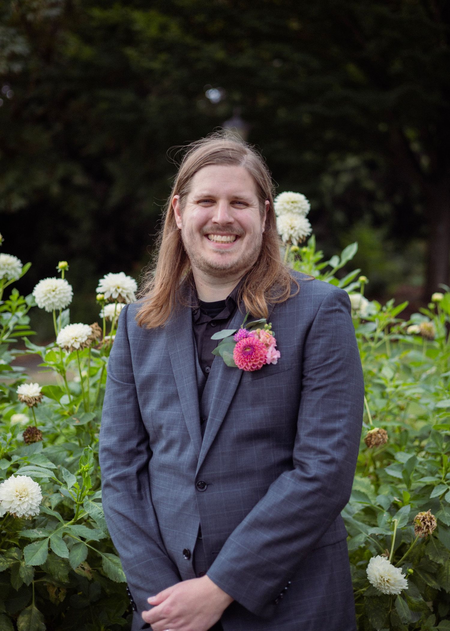 Smiling man with long hair wearing a gray suit and pink boutonniere standing in front of white dahlia flowers outdoors.
