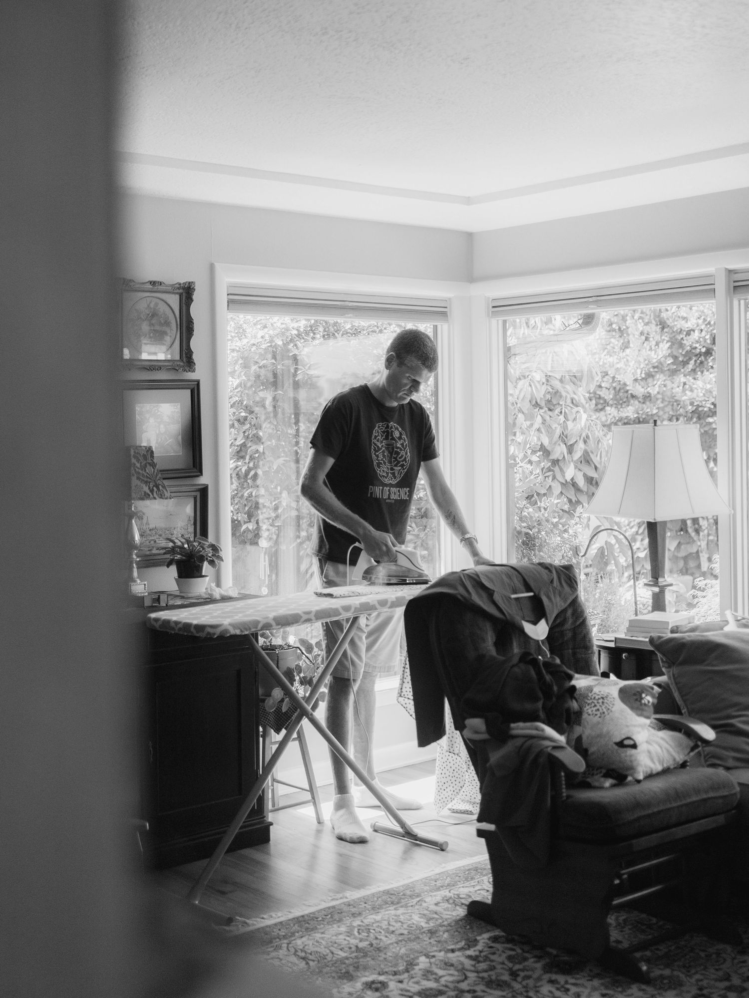 Man ironing clothes on an ironing board in a bright living room, black and white photo.