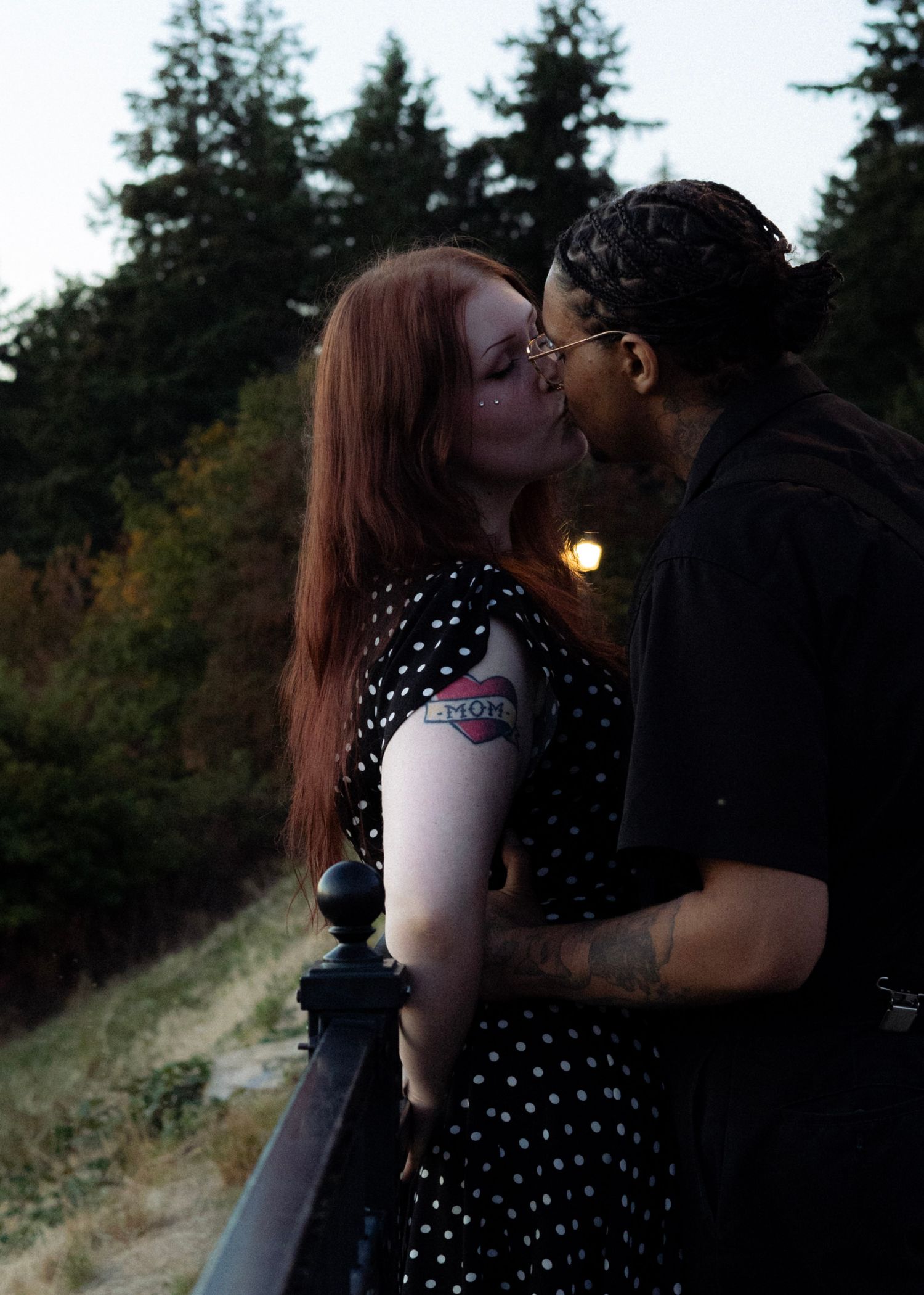 Couple kissing at dusk on a balcony, woman with red hair in polka dot dress with visible tattoo.