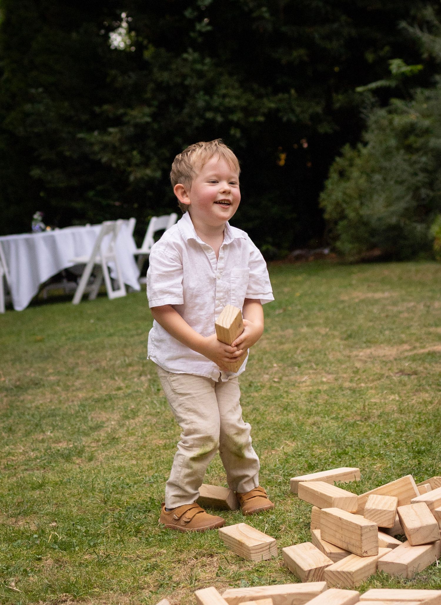 A laughing toddler boy in a white shirt plays with giant Jenga blocks on a green lawn at an outdoor event.