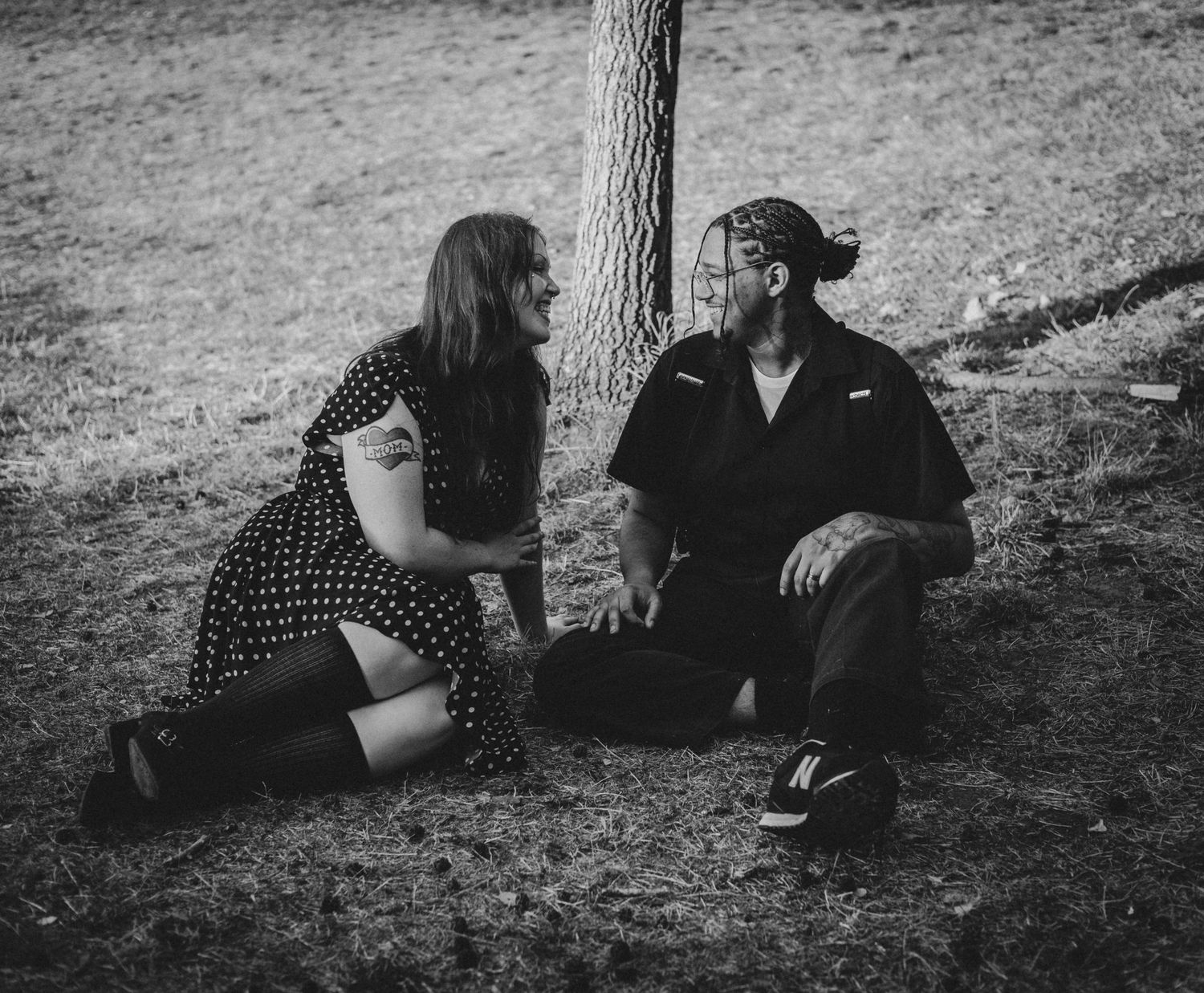Black and white photo of a couple sitting together on grass near a tree, sharing a conversation outdoors.