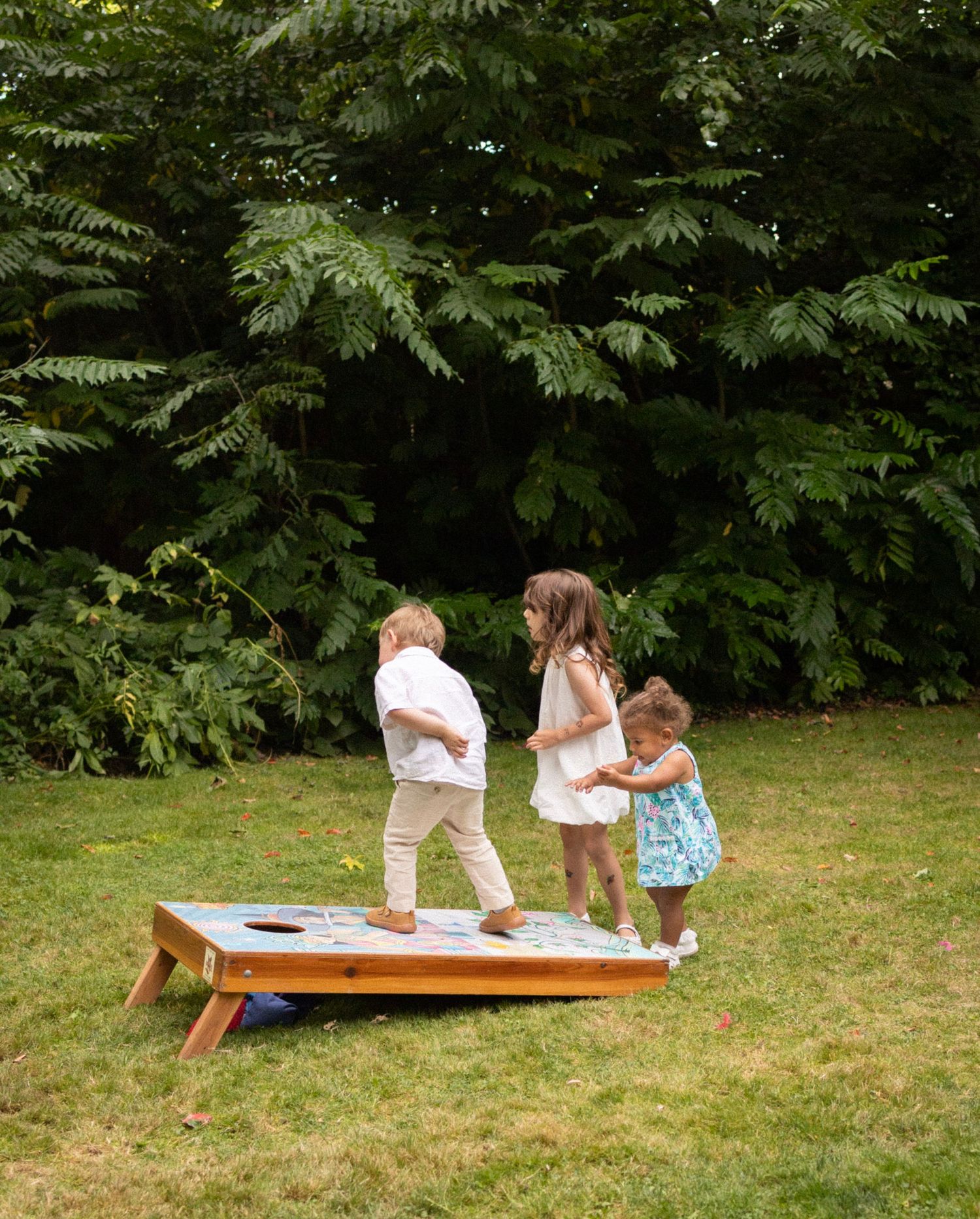 Three young children play on cornhole board game on green lawn at wedding.