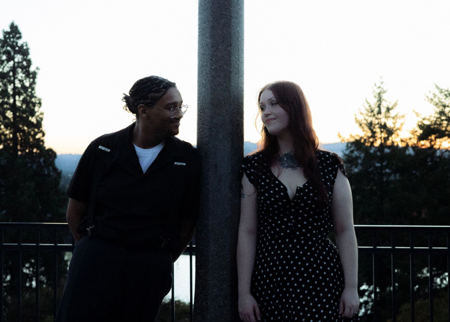 Couple standing apart by a pole at sunset, woman in polka dot dress and man in black shirt outdoors.