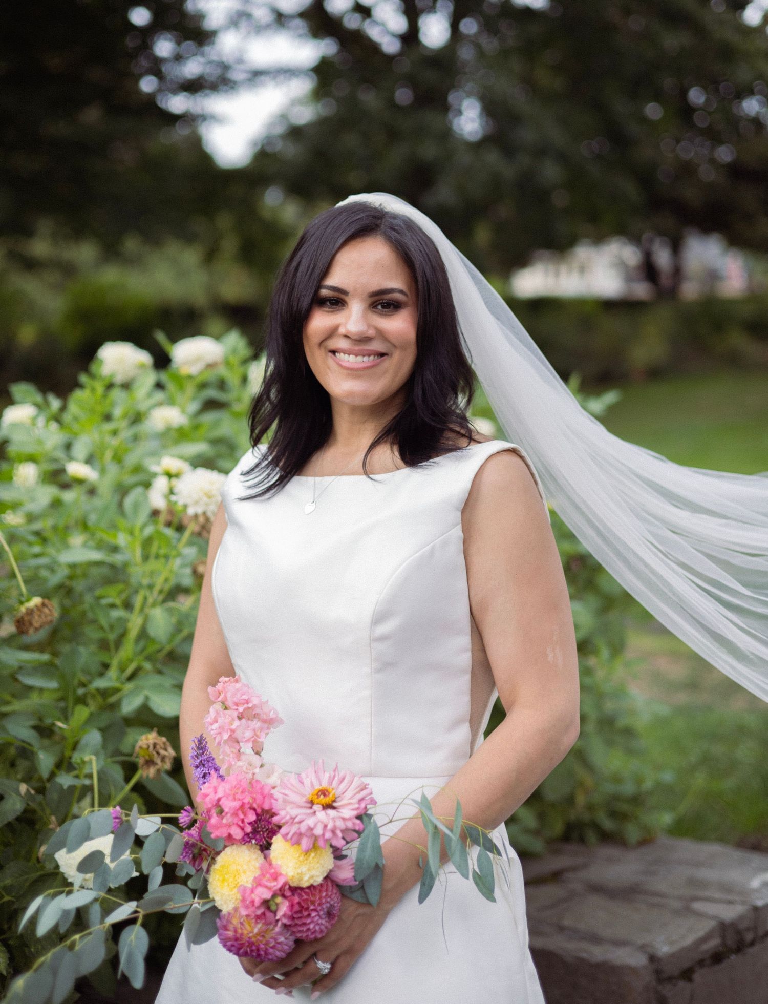 Smiling bride in white sleeveless gown and veil holding colorful pink bouquet in lush garden setting.
