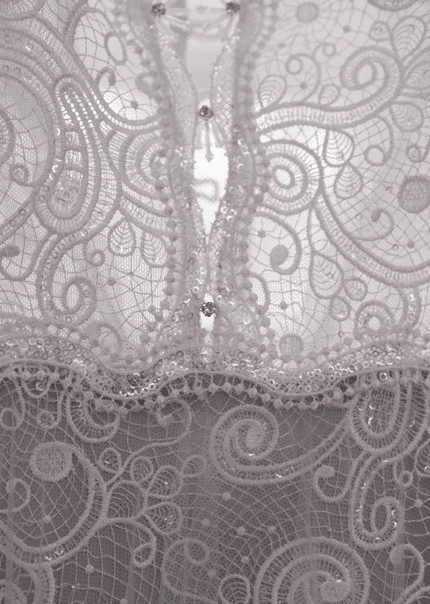 Close-up of delicate white lace fabric with intricate swirl patterns, beaded trim, and sparkling crystal embellishments.