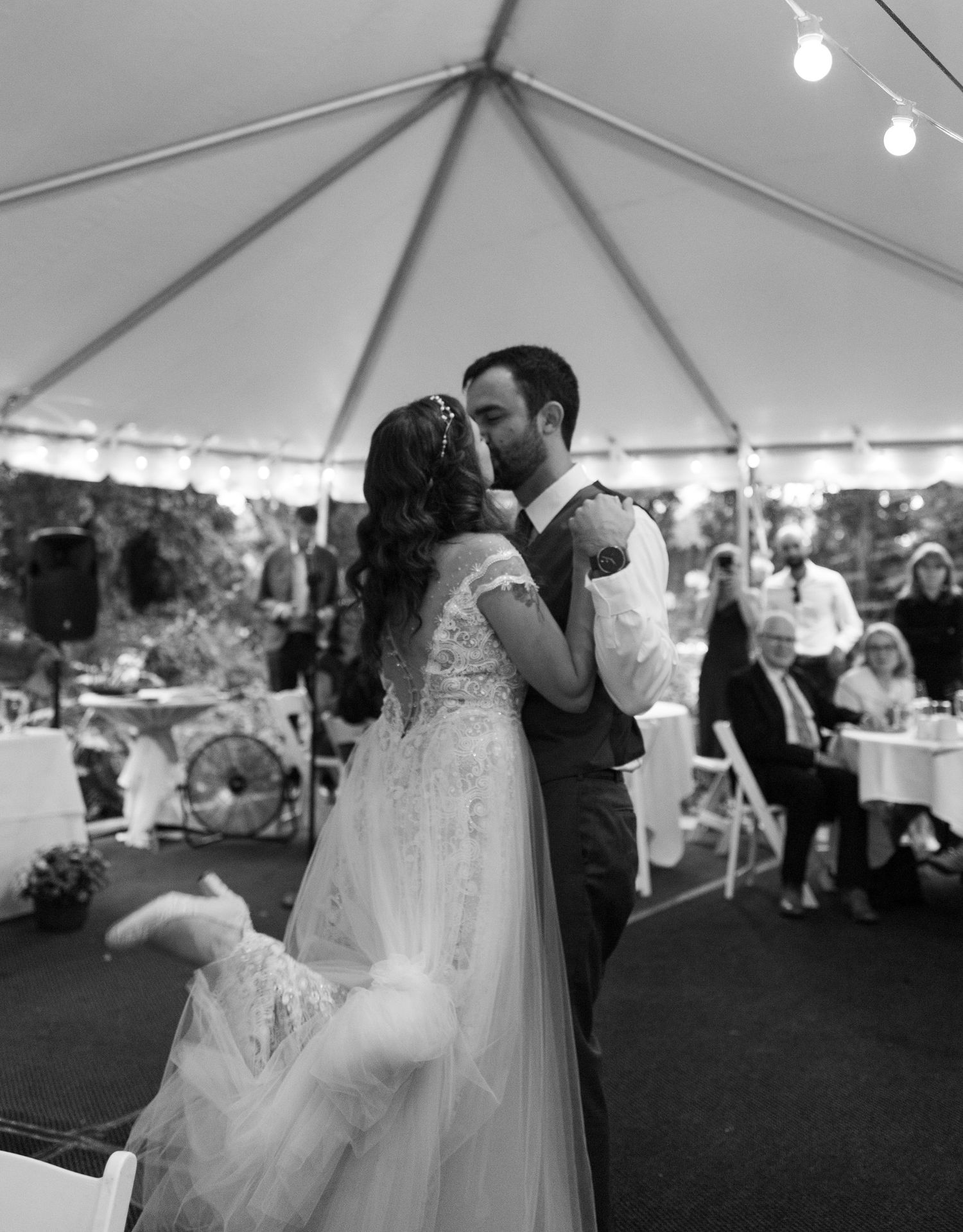 Black and white photo of bride and groom sharing first dance kiss under elegant tent at outdoor wedding reception.