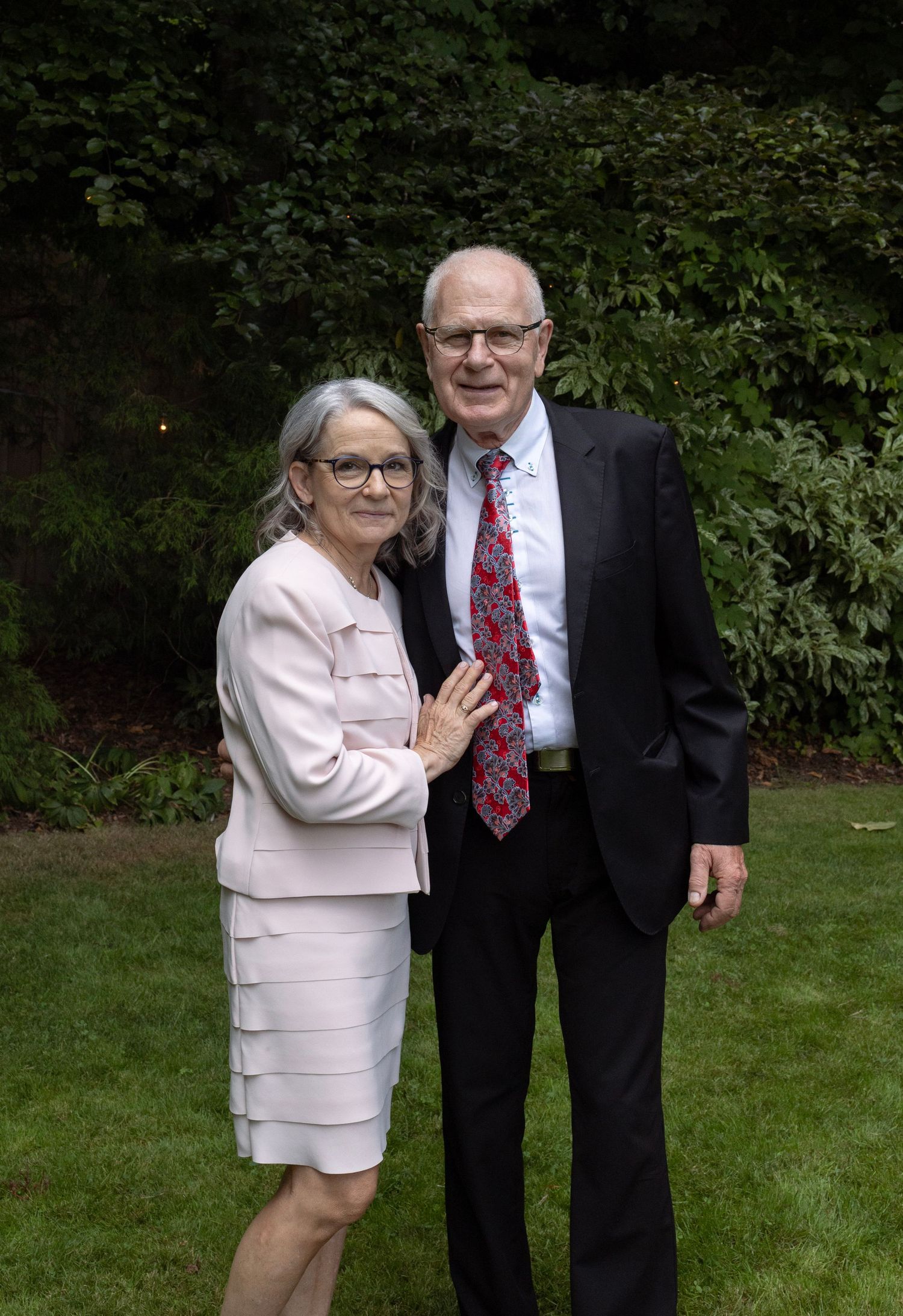 An elegant older couple poses together in a lush green garden at a wedding celebration.
