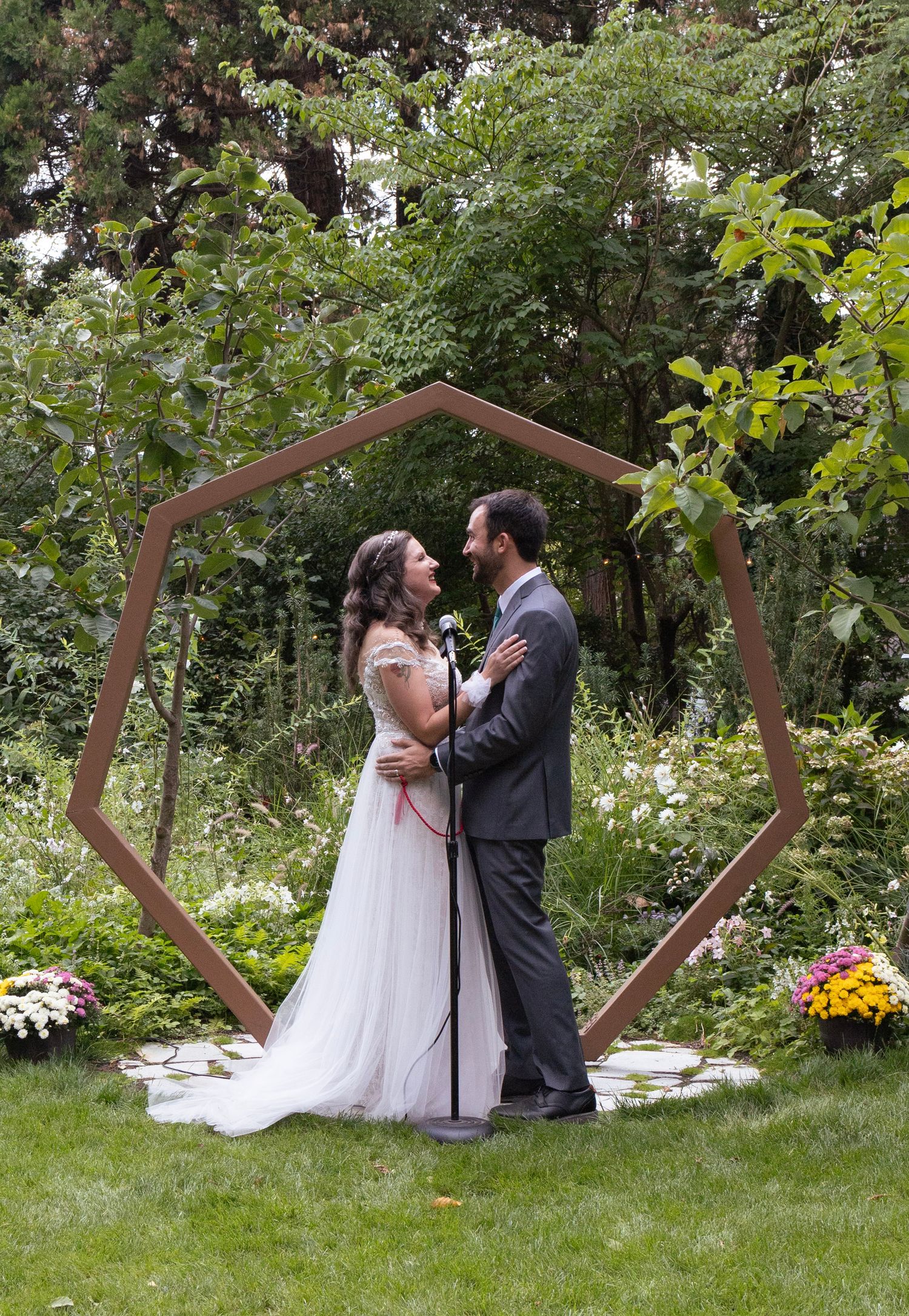 Bride and groom standing face to face under a geometric hexagonal wooden arch in a lush garden setting.