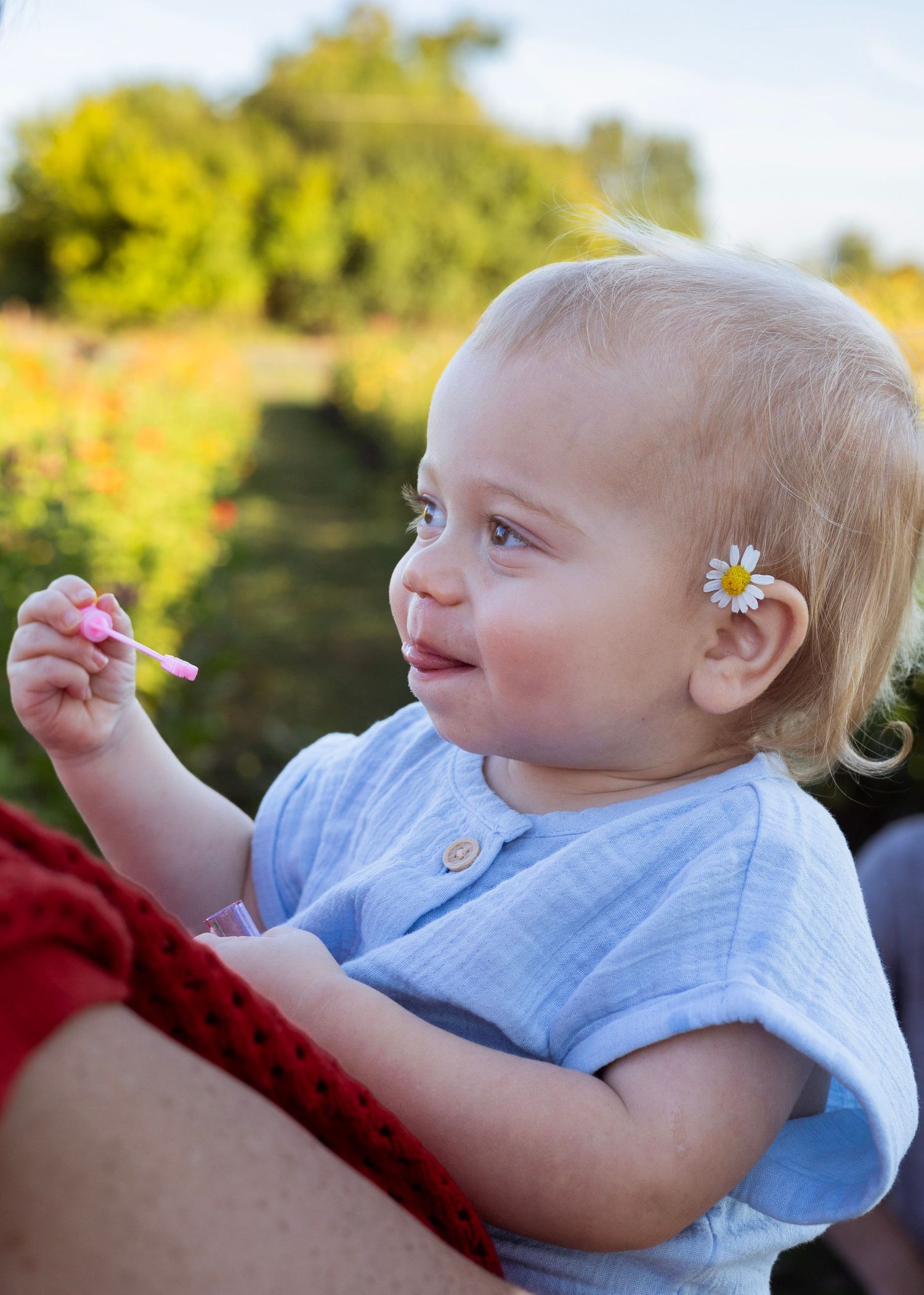 Smiling baby with daisy hair clip holds a pink toy outdoors in a sunny garden setting.