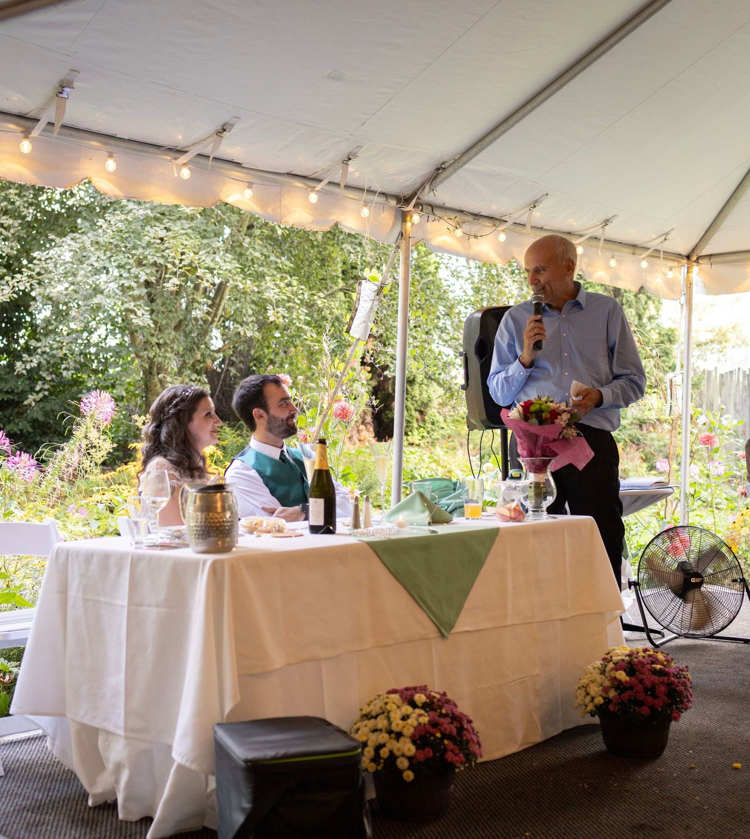 Father gives speech to bride and groom at floral decorated wedding reception table.