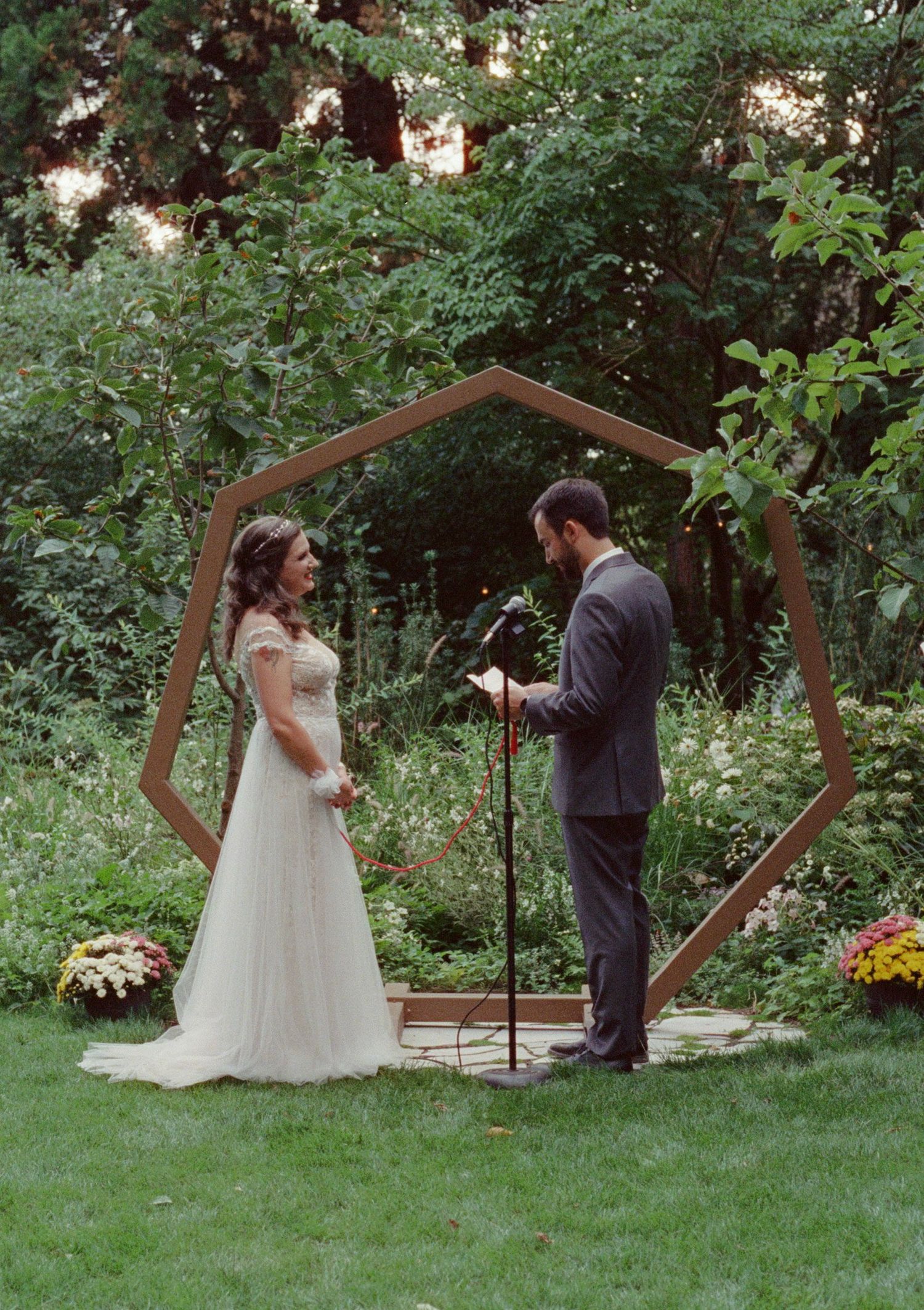 Couple exchanging vows outdoors under a geometric hexagonal wooden arch surrounded by lush garden greenery.
