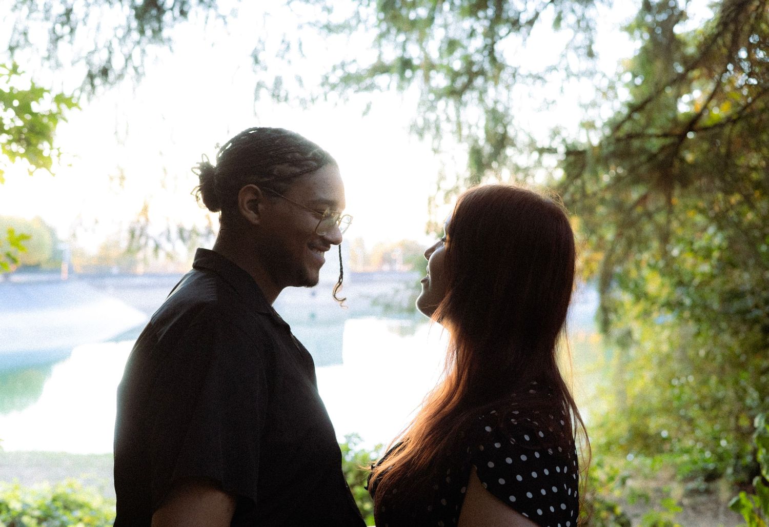 Couple sharing a romantic moment outdoors near a sunlit lake, surrounded by lush green trees.