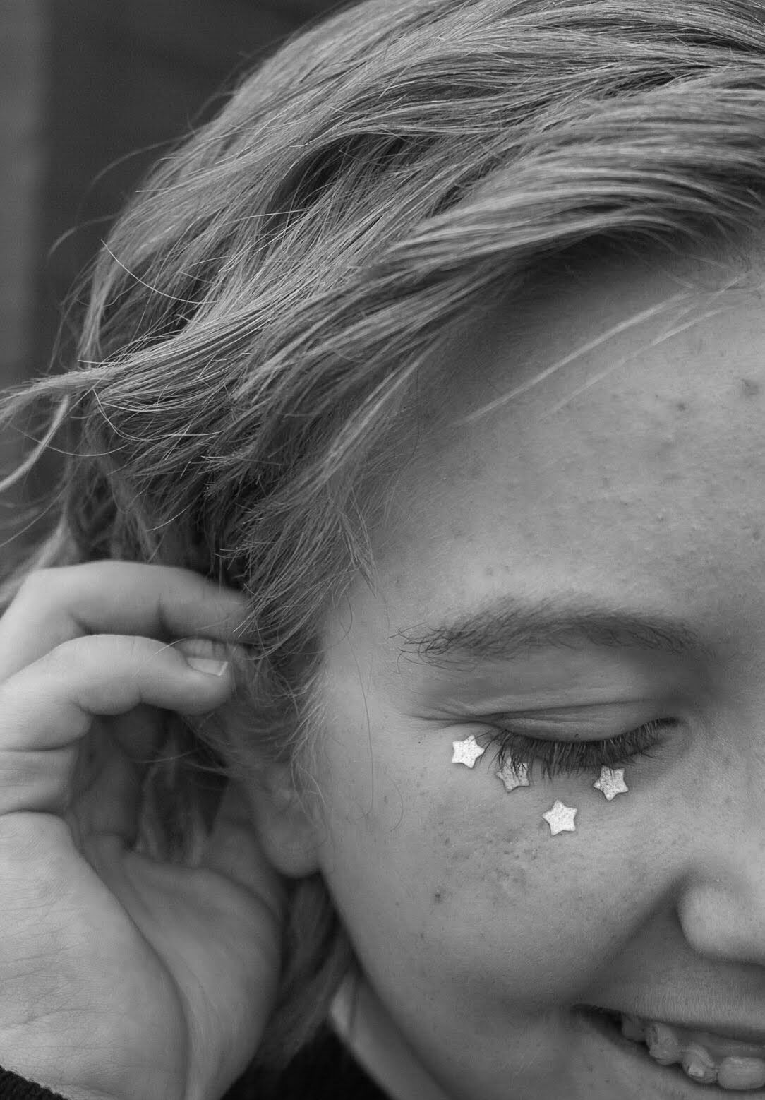 Black and white close-up of a smiling girl with star-shaped glitter beneath her eye.