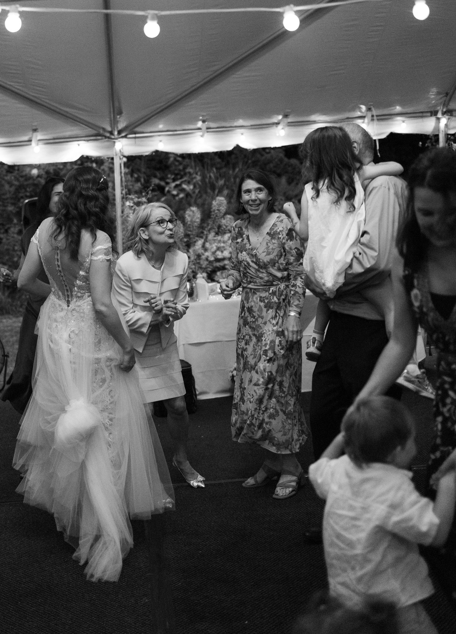 Black and white photo of wedding guests socializing under a tent, with bride in lace gown chatting with guests.