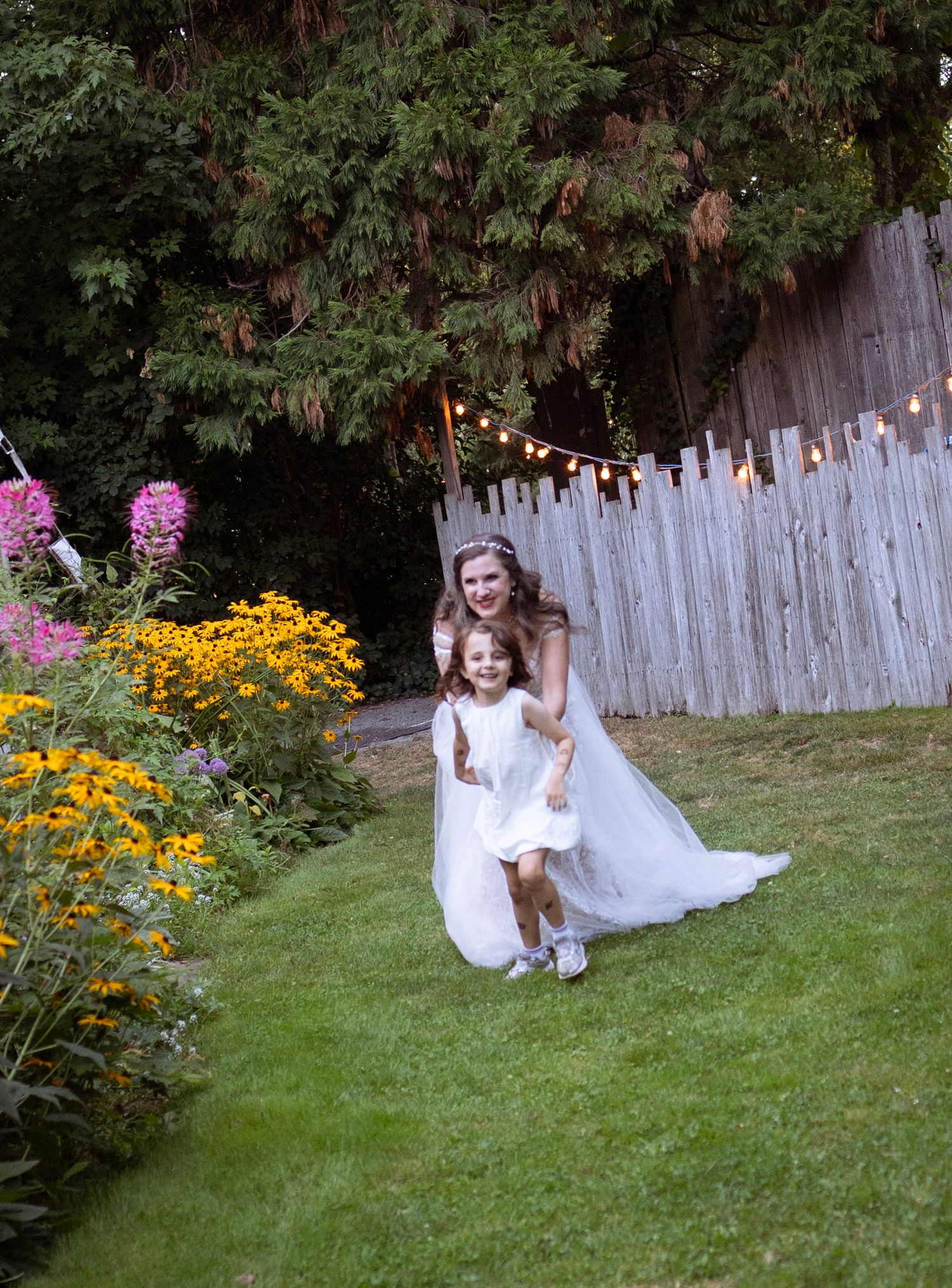 Bride in white gown running with flower girl through garden wedding venue with string lights.