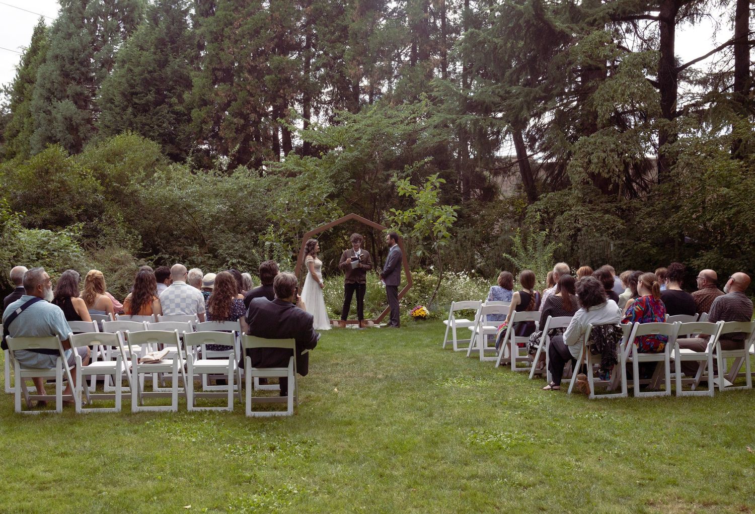 Outdoor wedding ceremony on green lawn with guests seated in white chairs facing couple at floral arch surrounded by tall trees.