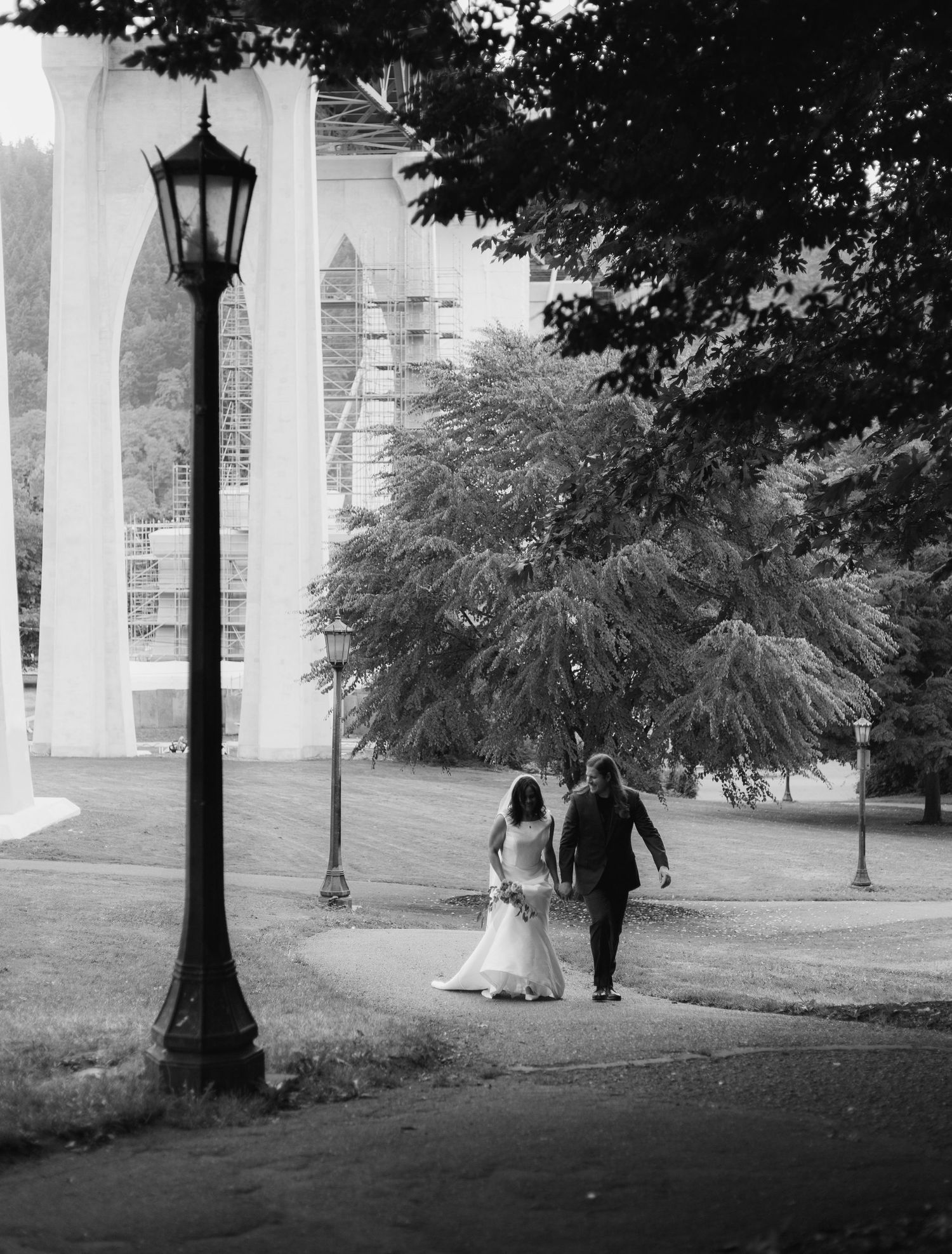 Black and white photo of a bride and groom walking hand in hand near a lamp post and bridge in a park.