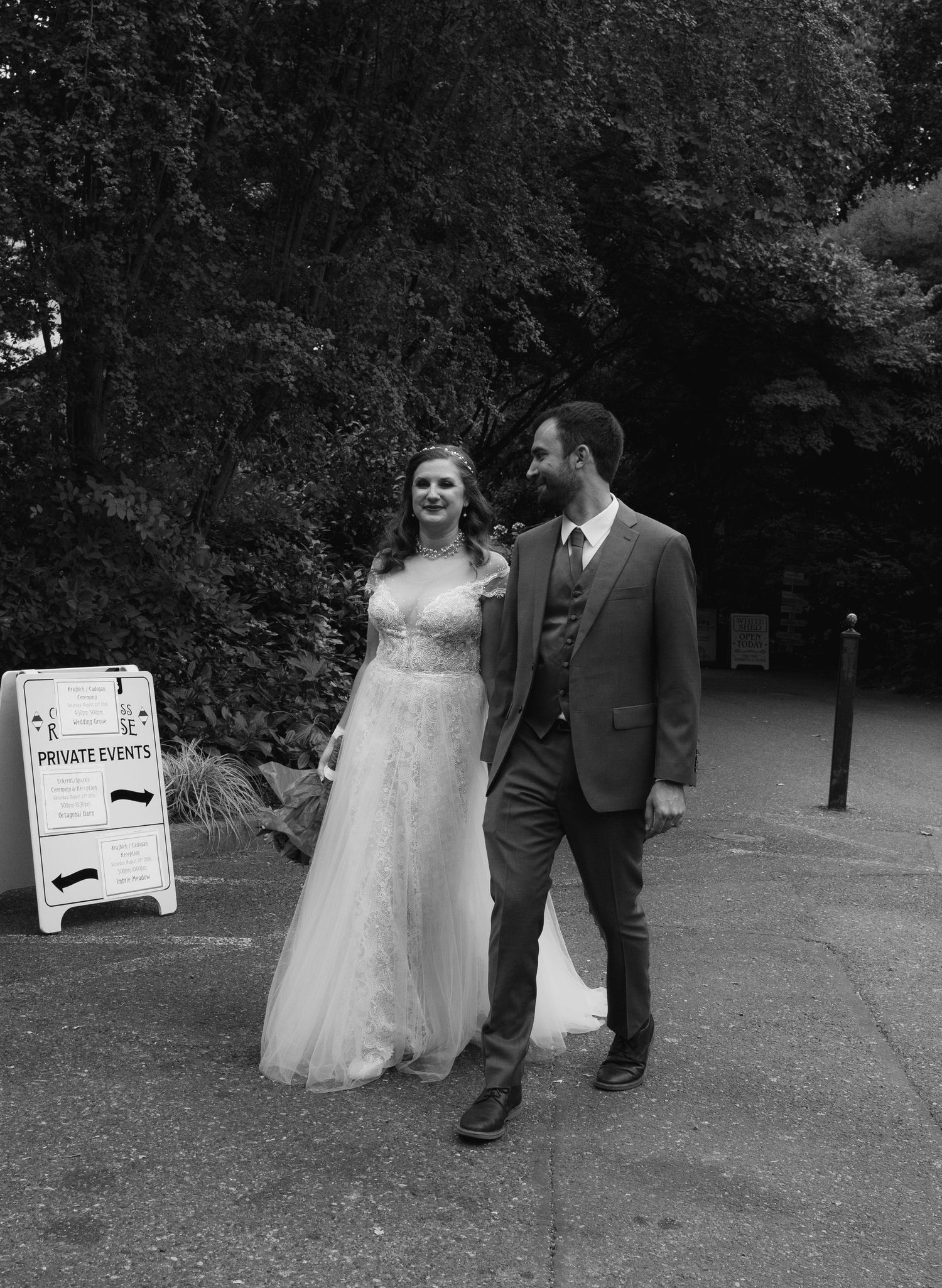 Black and white photo of a smiling bride in a white gown and groom in a suit walking together outdoors at a private event.