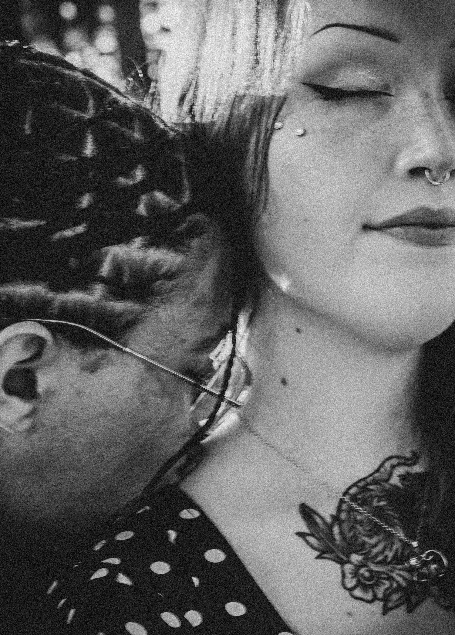 Black and white photo of a woman with nose piercing, face tattoos, and chest tattoo beside another person.