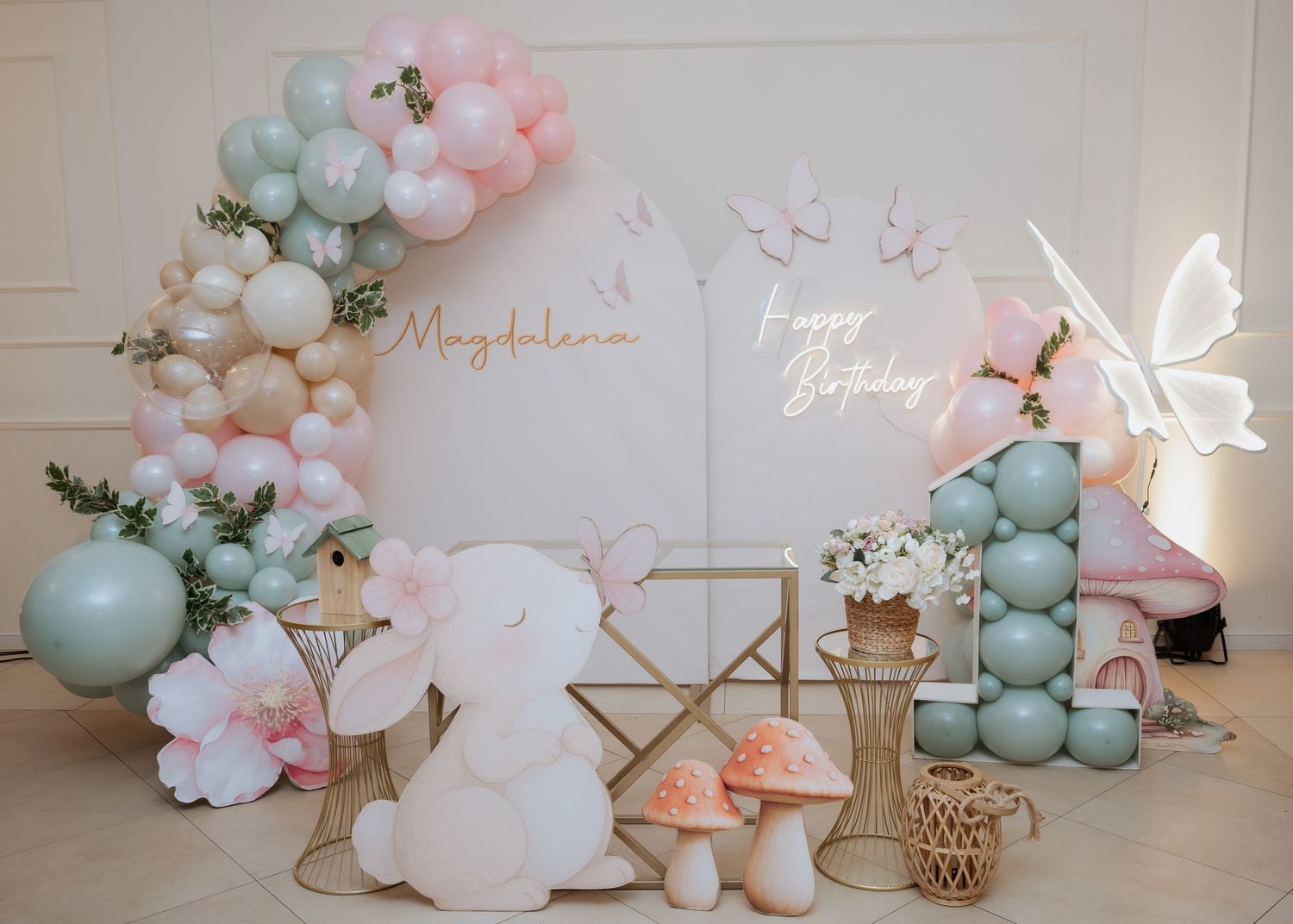 Whimsical first birthday party decor featuring pastel balloons, a bunny figurine, mushrooms, and a Happy Birthday neon sign.