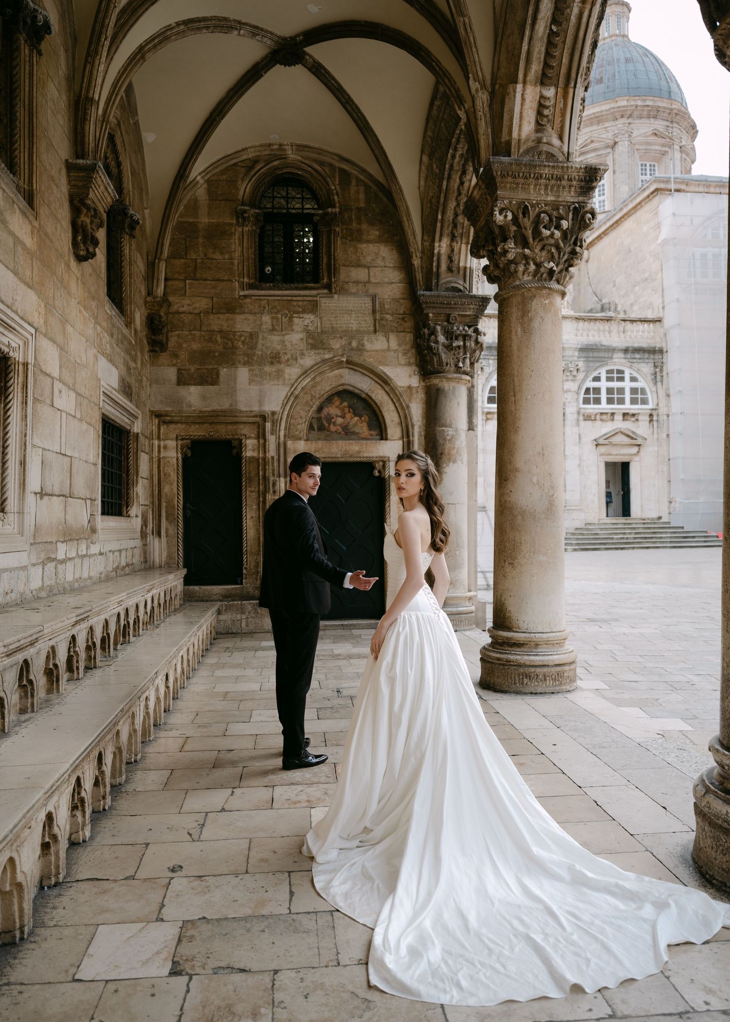 Bride and groom pose under stone arches of historic Dubrovnik building in elegant wedding attire.