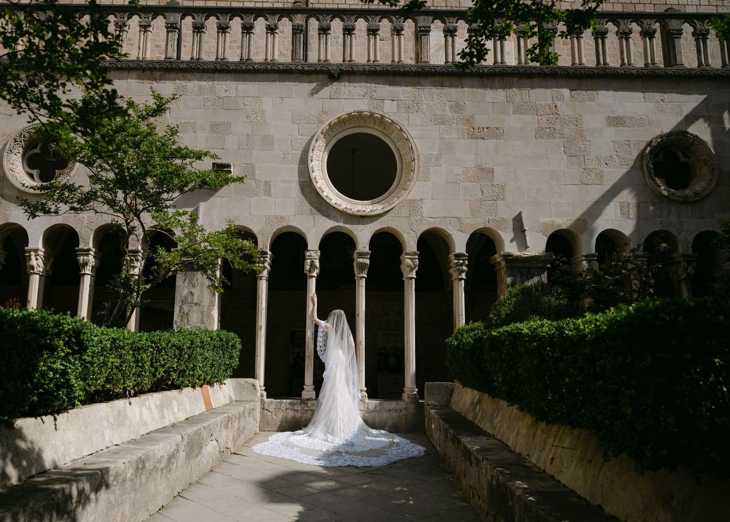 Bride in flowing white gown with cathedral train stands in sunlit Romanesque cloister with arched colonnade.
