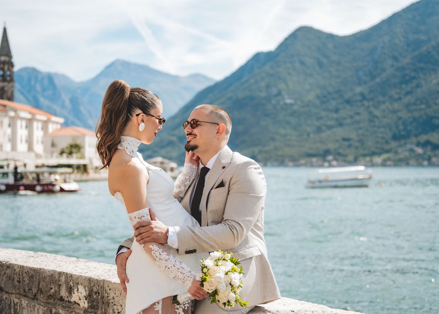 Bride and groom sharing intimate moment seated on stone wall with Bay of Kotor Montenegro backdrop.