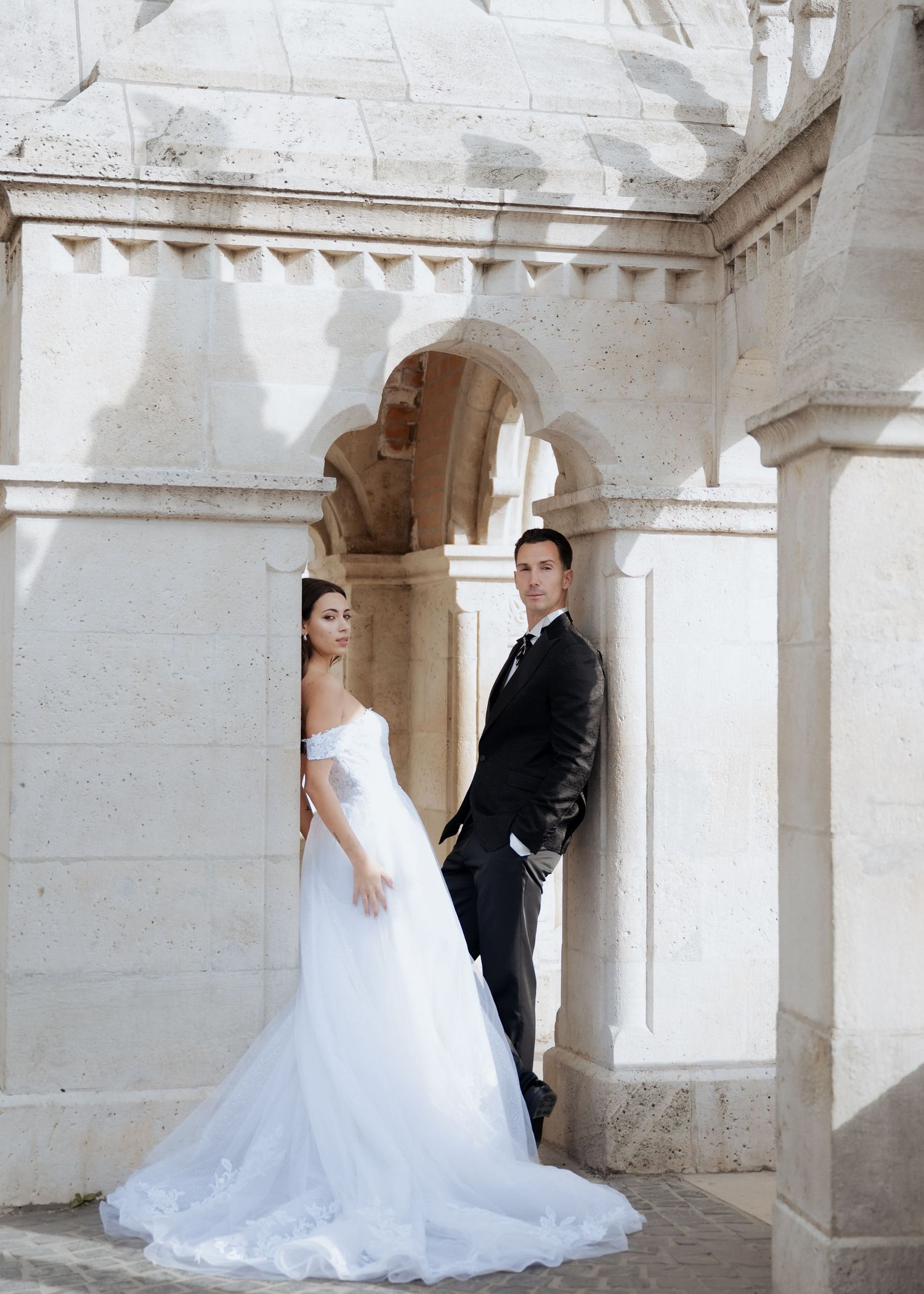 Bride and groom posing elegantly beneath stone arches of a historic European castle venue.