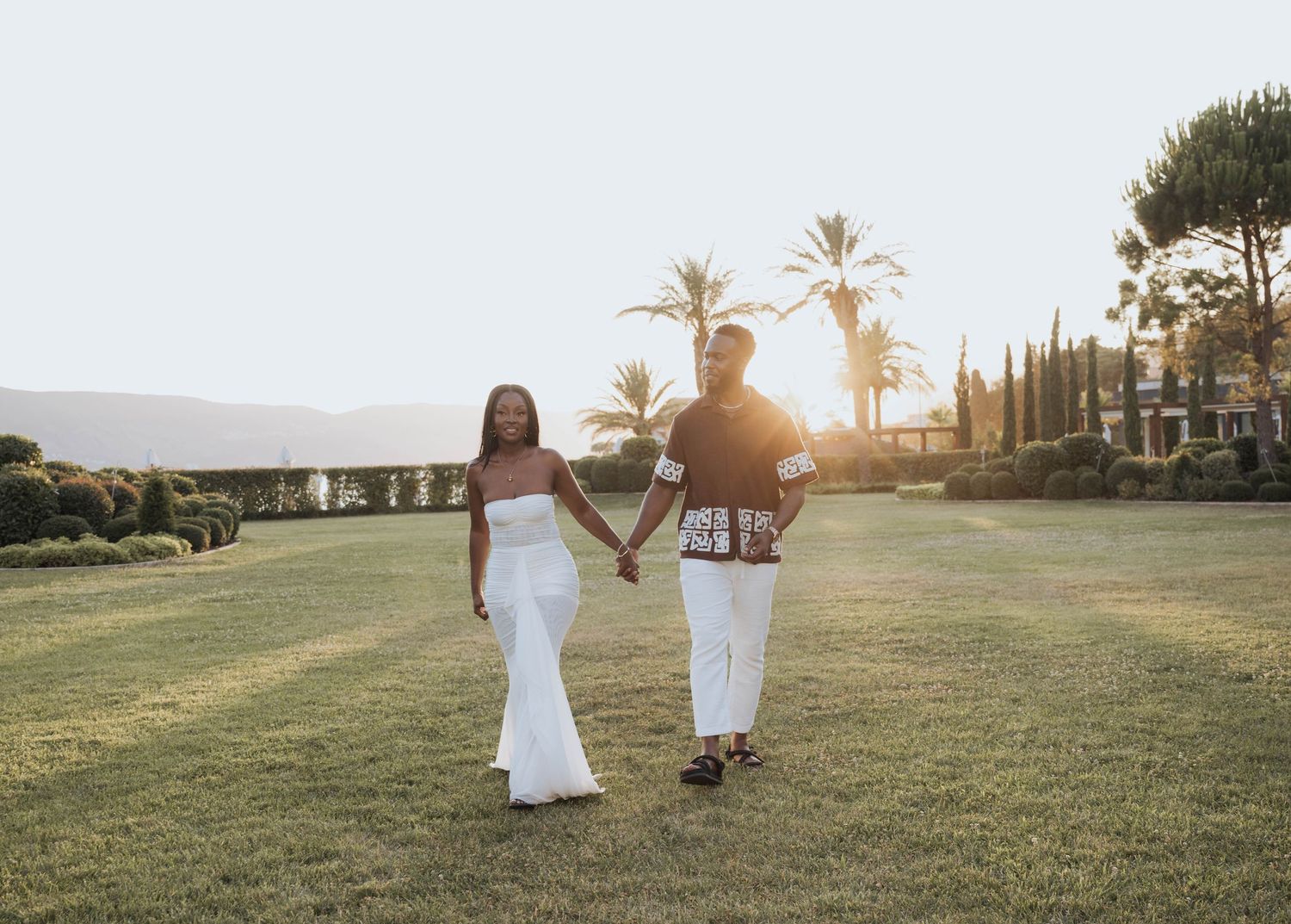 A newly engaged couple walking hand in hand across a green lawn at golden sunset.