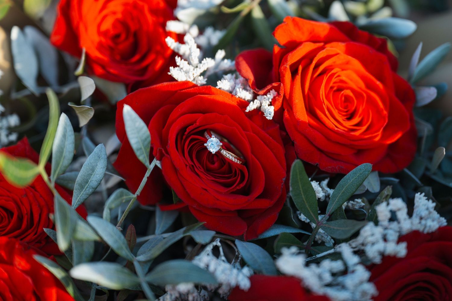 Close-up of a diamond engagement ring resting among vibrant red and orange roses in a bridal bouquet.