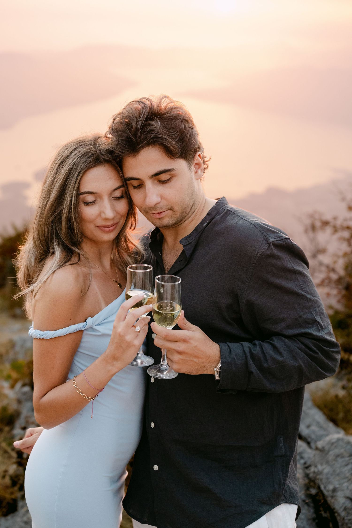 Couple toasting champagne glasses together at golden sunset with scenic mountain and lake backdrop.