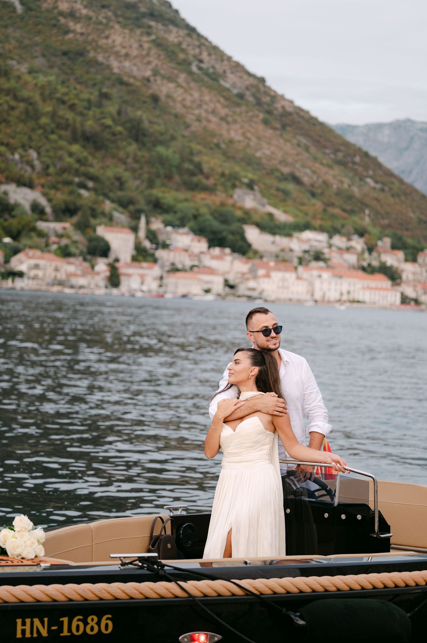 Wedding couple embracing on a boat with picturesque Perast village and Bay of Kotor in Montenegro background.