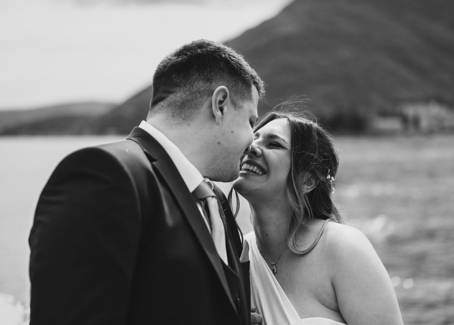 Laughing bride and groom share a joyful moment outdoors with a mountain lake backdrop.