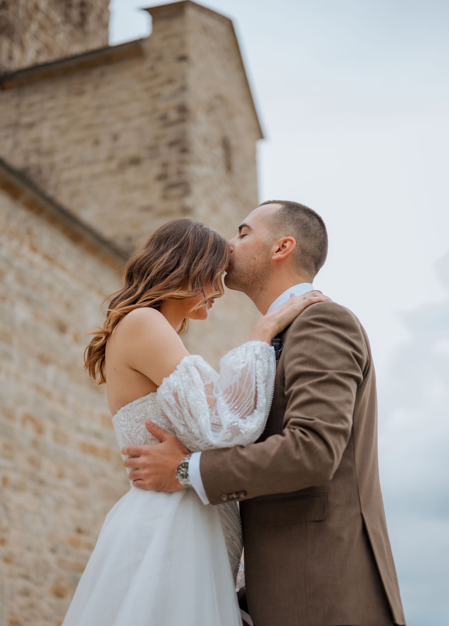 Groom in brown suit kissing bride in off-shoulder lace wedding dress by stone church.