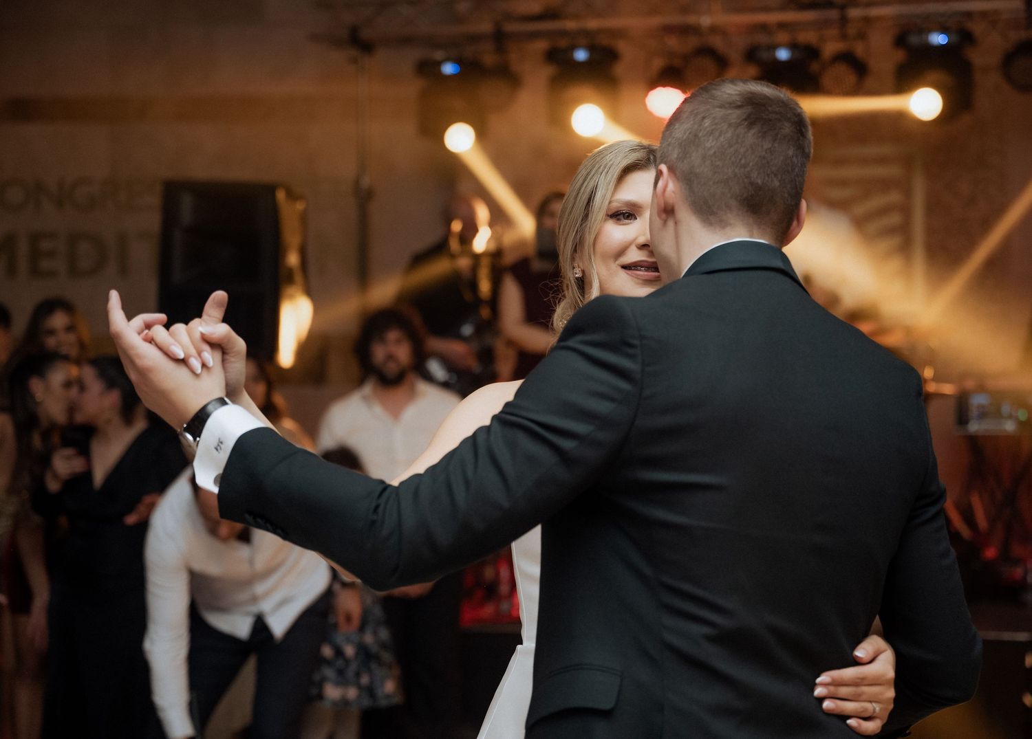 Bride and groom dancing closely at wedding reception with warm stage lighting and guests in background.